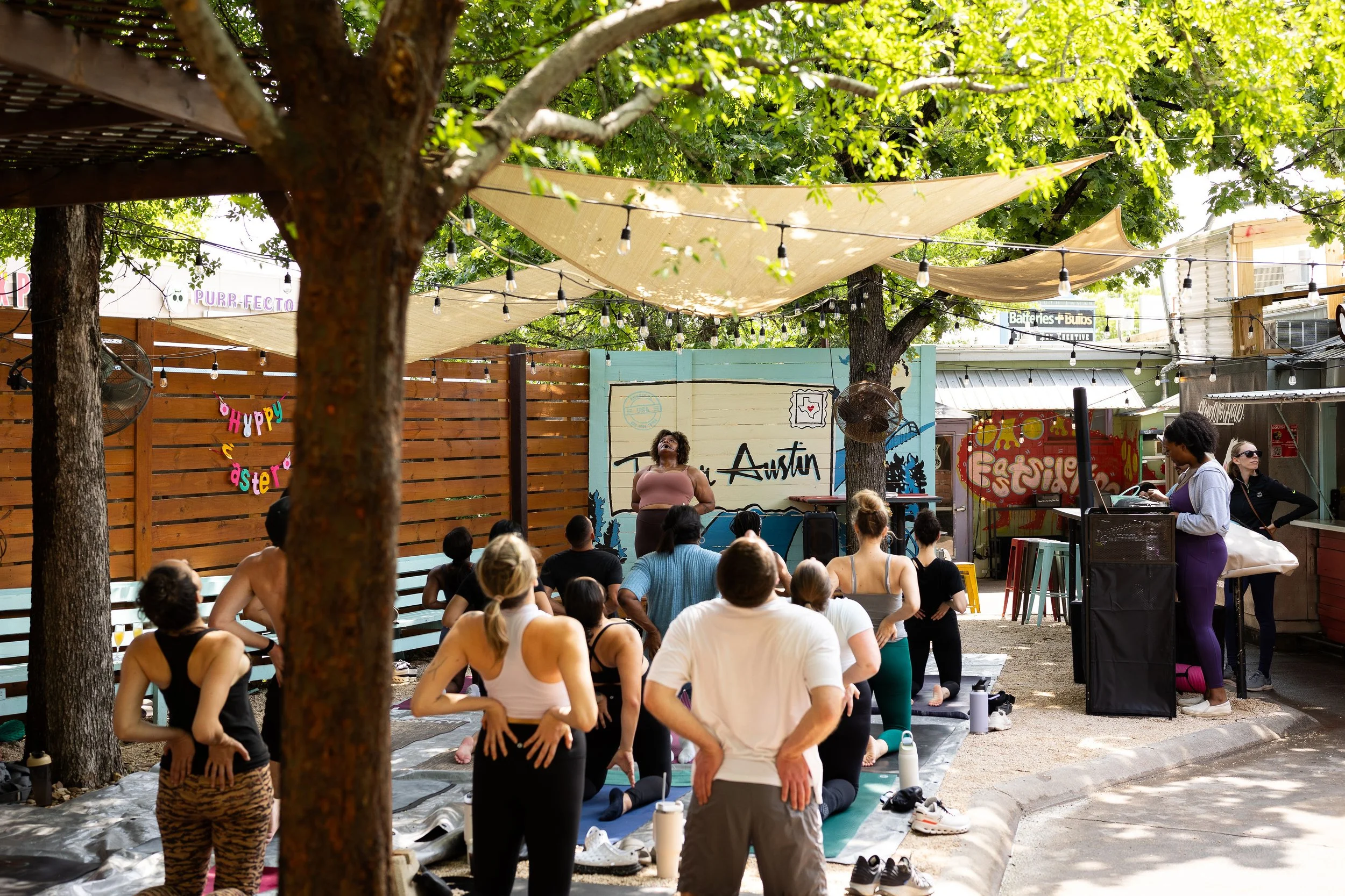 Group of people participating in outdoor yoga class under trees and string lights in a backyard or park, with a woman instructing at the front.