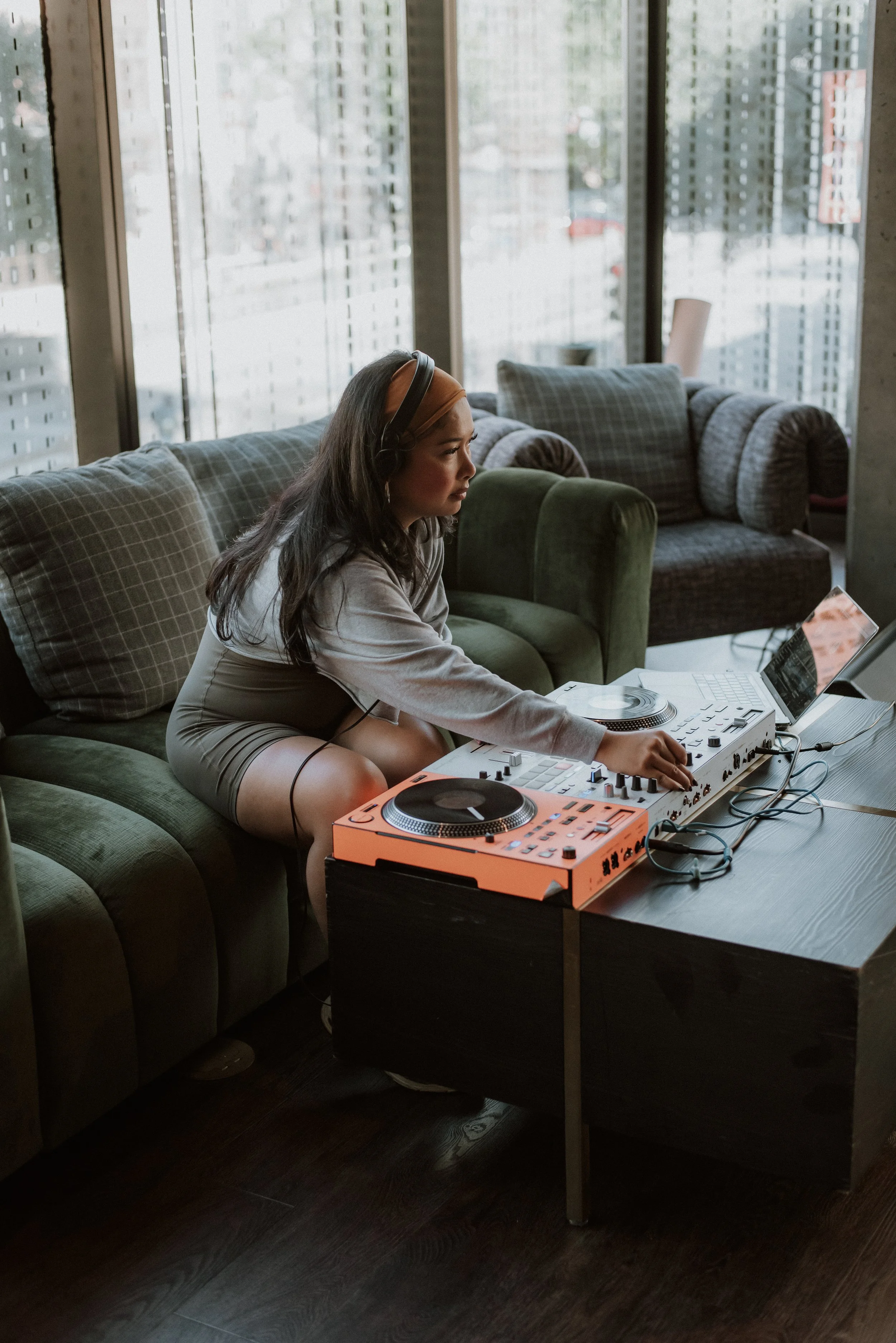 A young woman wearing headphones is DJing with a laptop and DJ controller on a table inside a modern room with large windows and sofas.
