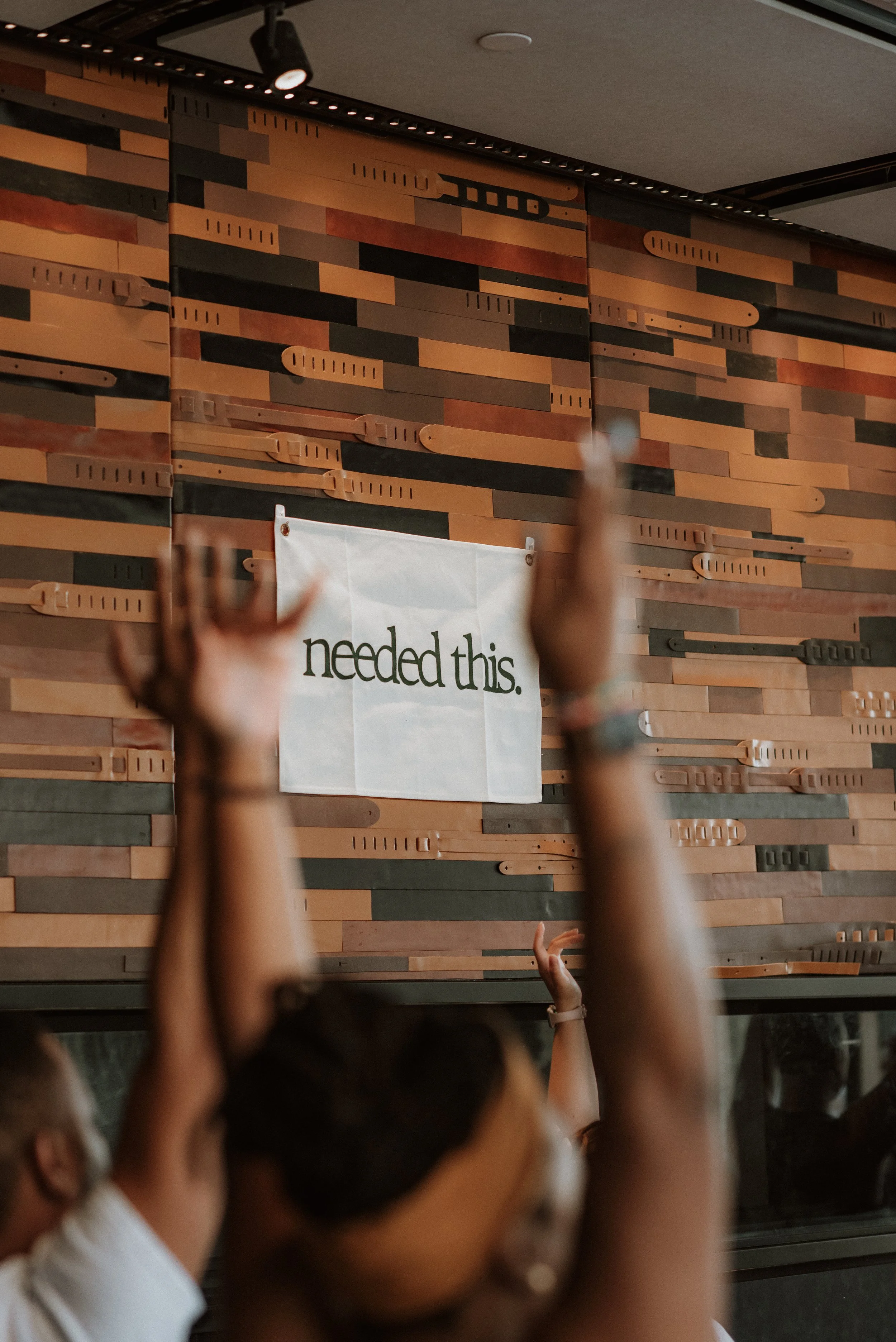People raising their hands in front of a wall with a sign that reads 'needed this' in a cafe or meeting space.