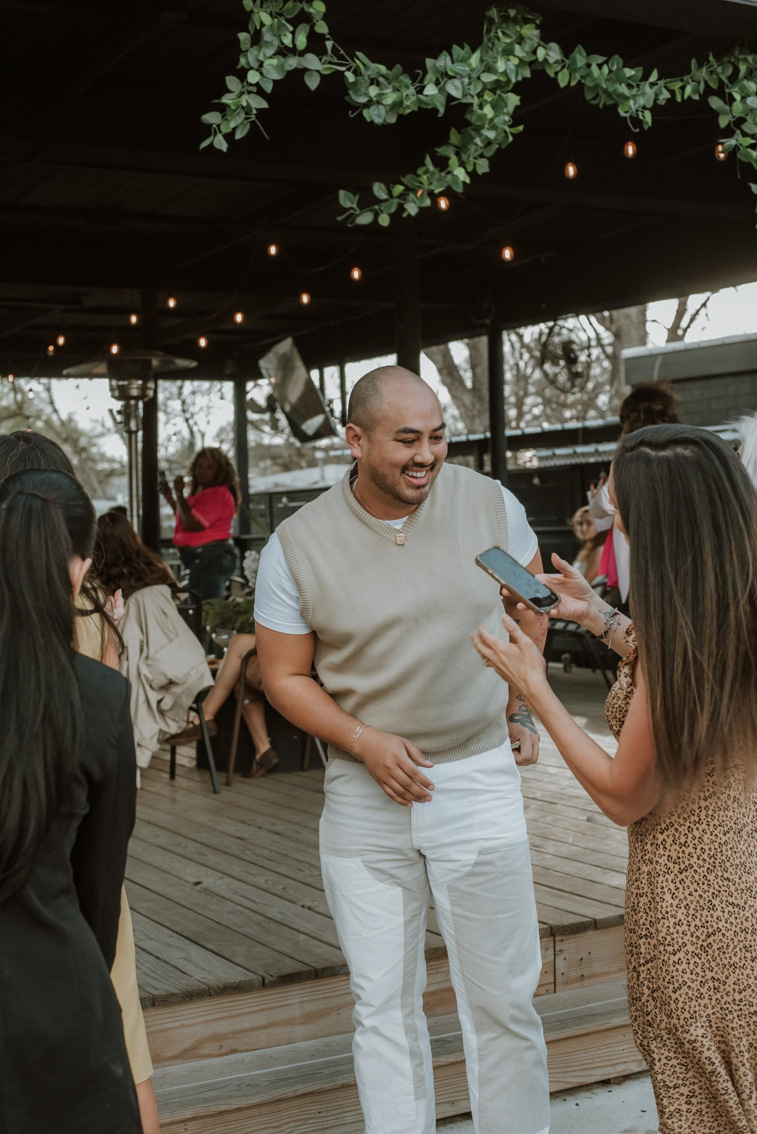 A group of people are gathered at an outdoor event, with a man in the center smiling and talking to a woman holding a smartphone, while others look on. There are string lights hanging and trees in the background.