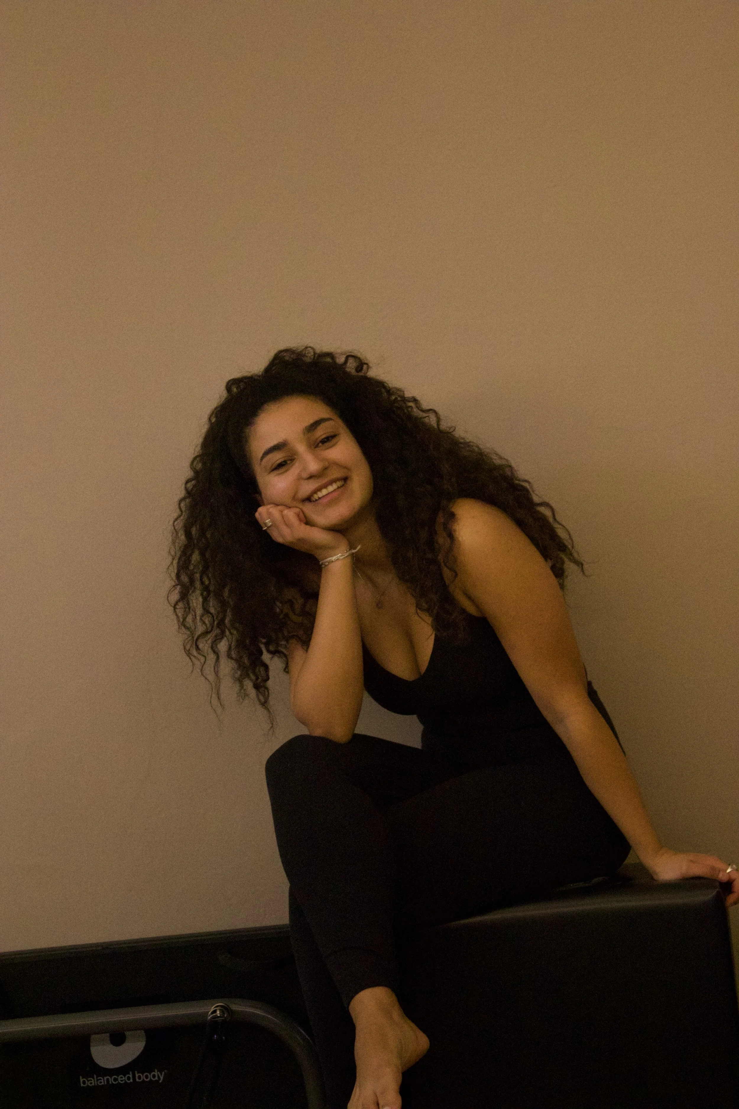 A smiling woman with curly hair, wearing a black tank top and black pants, sitting on a piece of exercise equipment against a beige wall.
