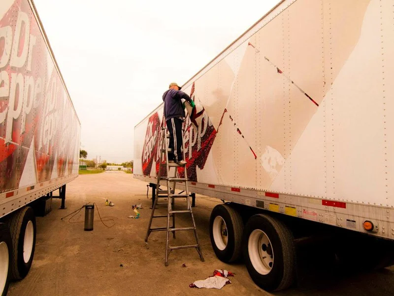 Person on a ladder applying or removing a large advertisement wrap on a semi-truck trailer.