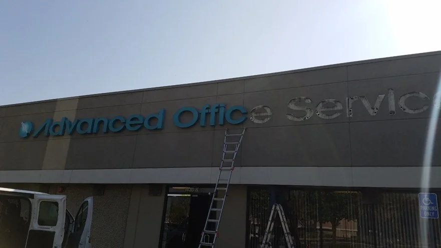 Sign reading 'Advanced Office Service' on the side of a building, with a ladder leaning against the sign, and a white vehicle parked nearby.