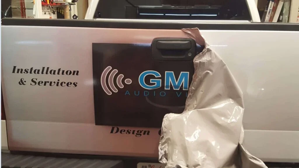 Close-up of a white vehicle door with a sign that reads 'Installation & Services' and 'Design', featuring a logo for GM Audio Video, with some brown paper wrapping partially covering the logo.