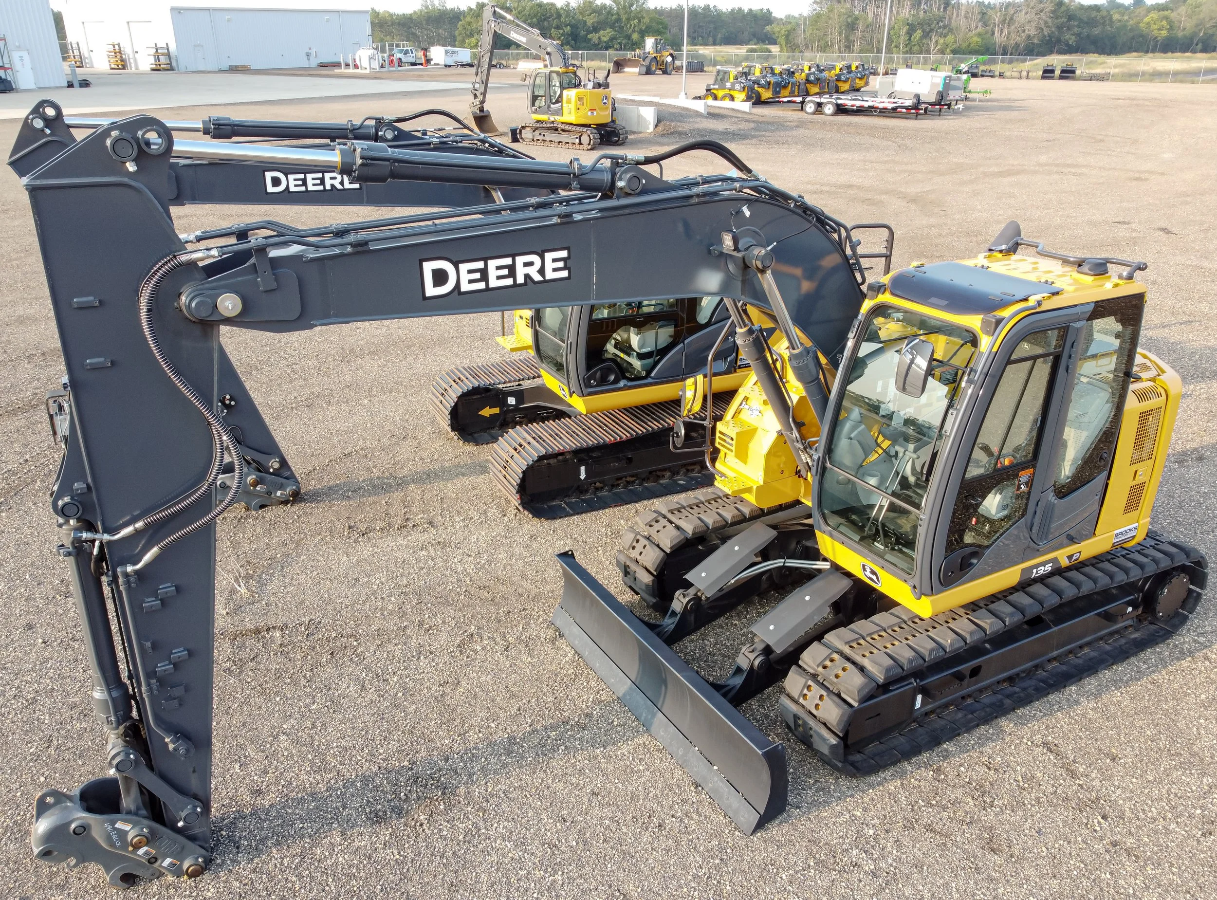 A Deere compact excavator with a black arm and yellow body, parked on gravel in an outdoor lot with other construction equipment and trailers in the background.