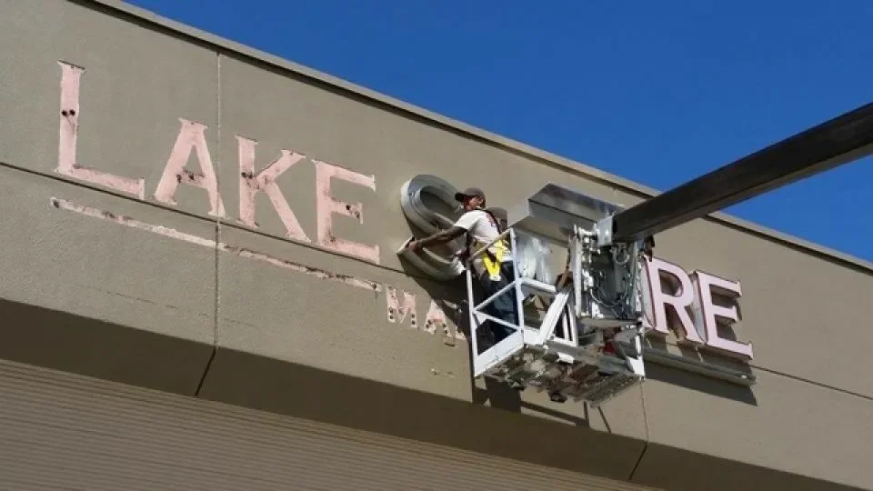 A worker in a cherry picker installing or repairing a sign that reads 'LAKE THE' on a building exterior against a blue sky.