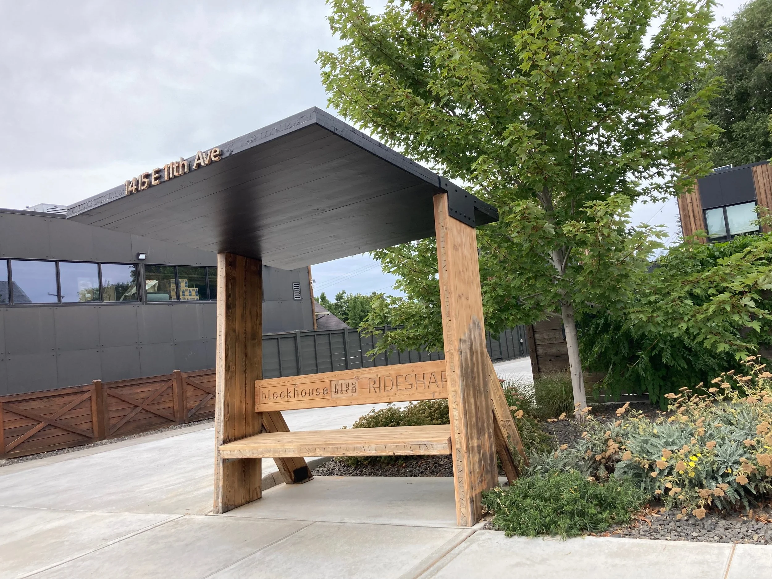 Wooden bus shelter with a black roof and the address 145E 11th Ave on top, surrounded by green trees and plants, with a building in the background.