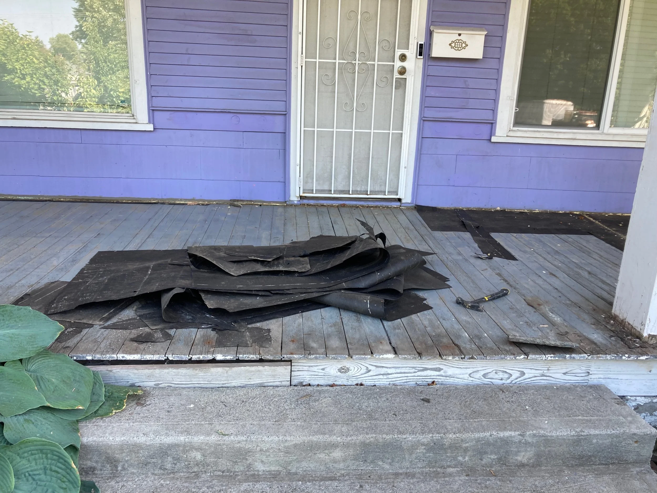 Front porch with purple house, white door with decorative security grille, mailbox, boarded-up area, pile of black roofing material, and a pair of roofing shears on the porch floor.