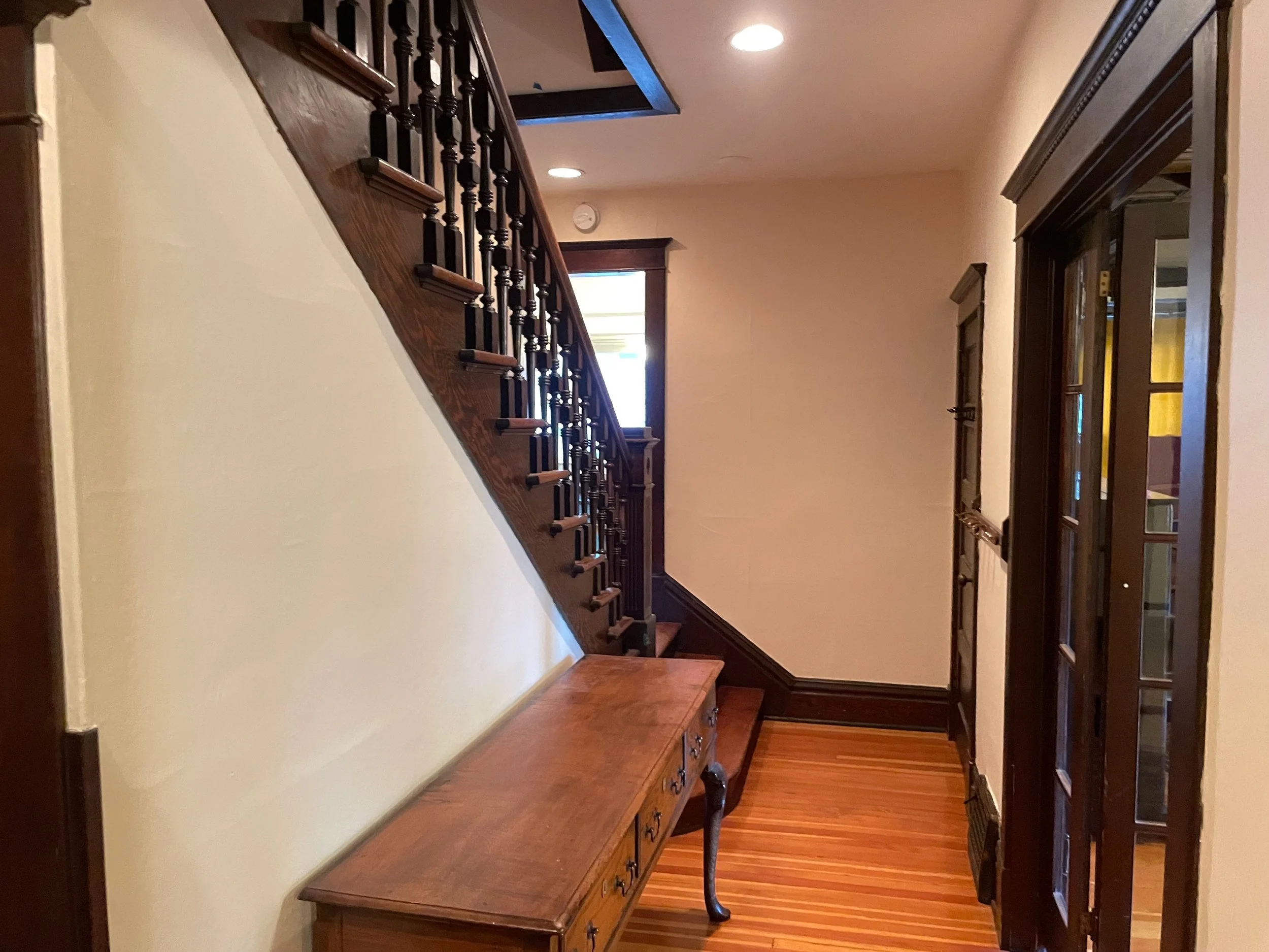 Interior view of a hallway with wooden staircase on the left, a wooden console table beneath it, and a door to the right. The hallway has beige walls, hardwood flooring, and trim with dark wood finish.
