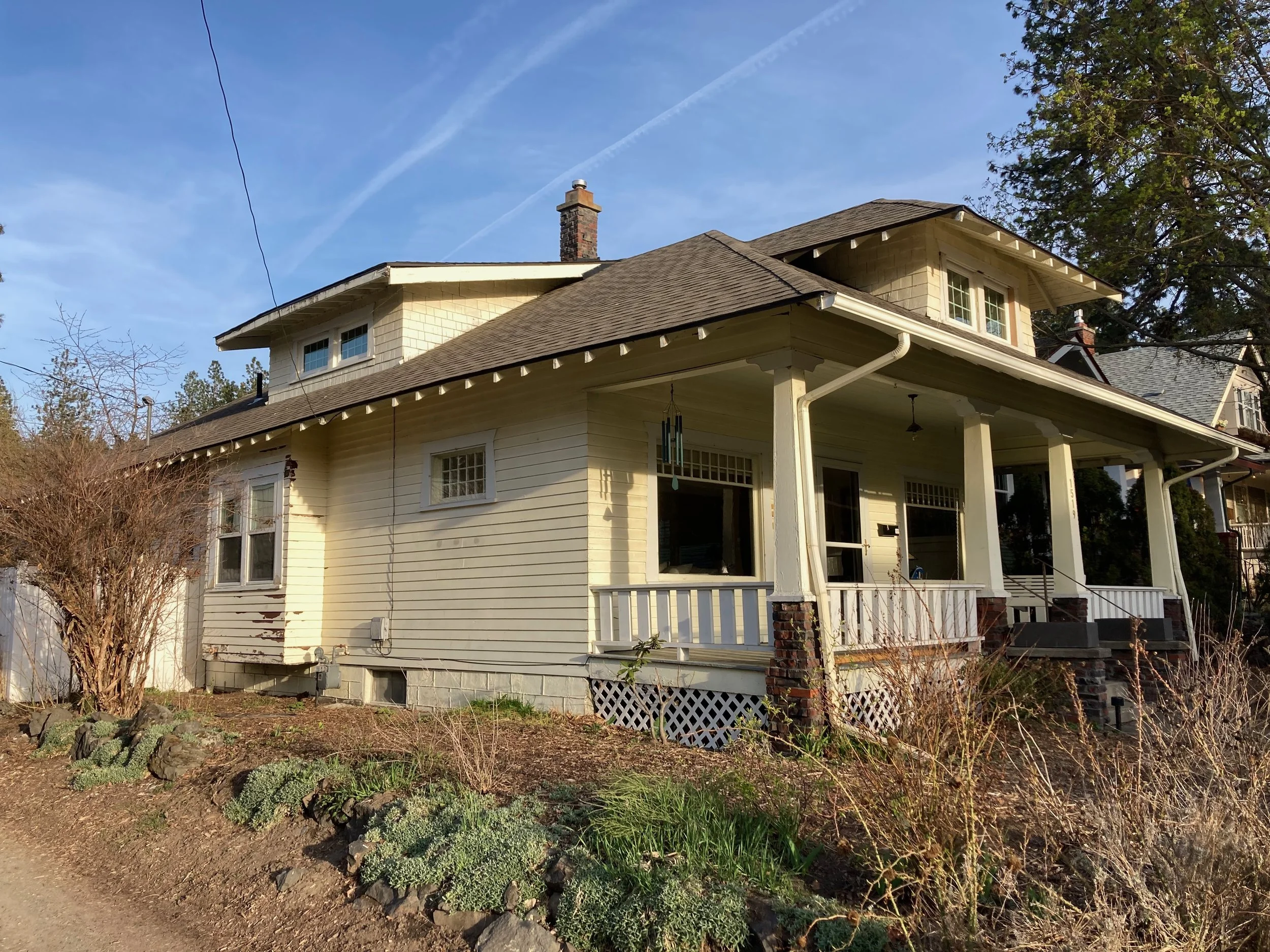 A two-story house with a porch, cream-colored siding, brick foundation, and a shingled roof. The house has multiple windows and a chimney, with some trees and bushes in the front yard. The sky is blue with visible contrails.