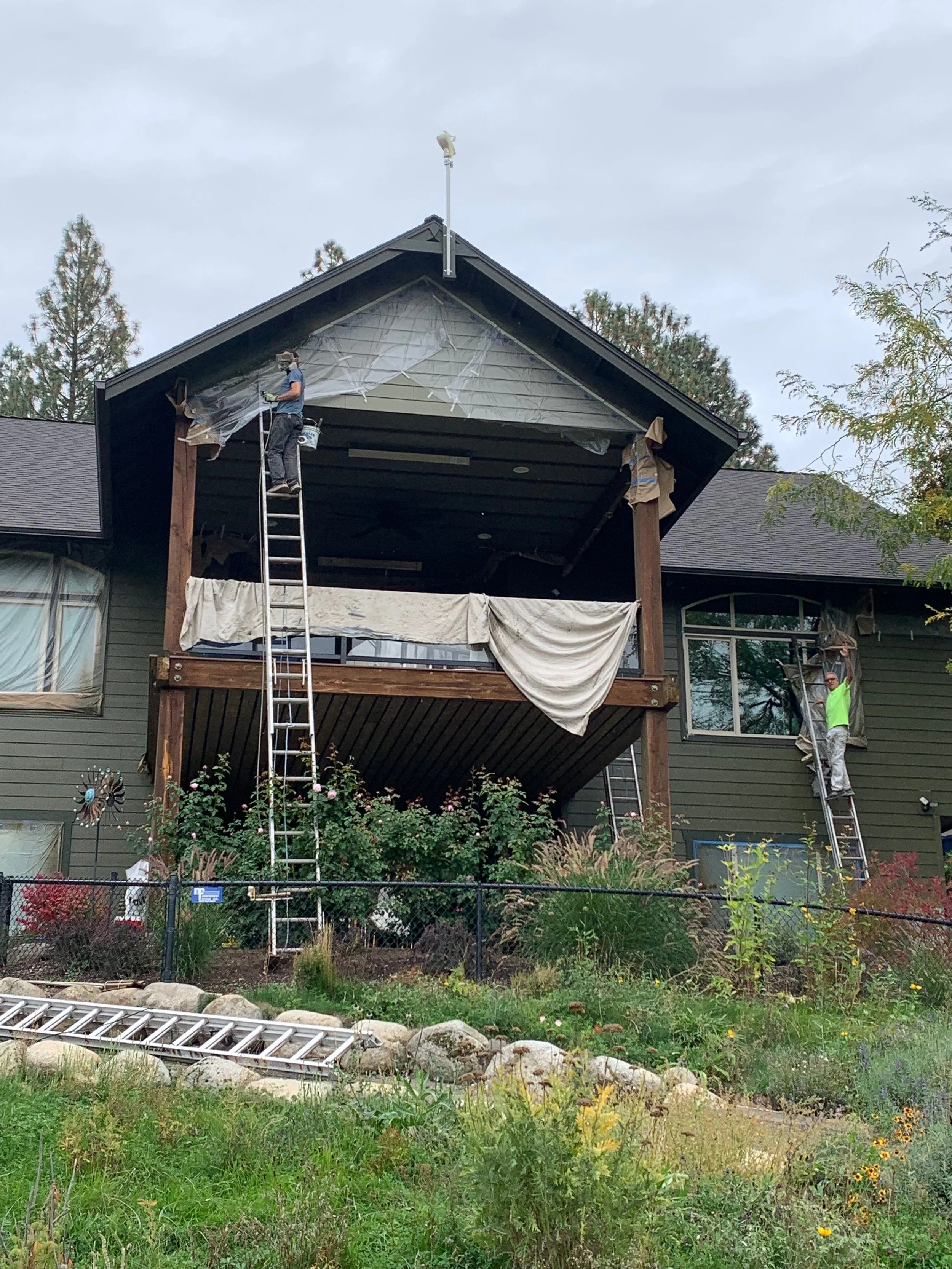 Two workers are painting and repairing the exterior of a house's second-story balcony and window during the daytime. One worker is on a ladder near the balcony, applying paint or repairs to the upper part of the house, while the other is on a ladder near the window, working on the house facade. The house has dark green siding, and the balcony is decorated with plants and flowers below.