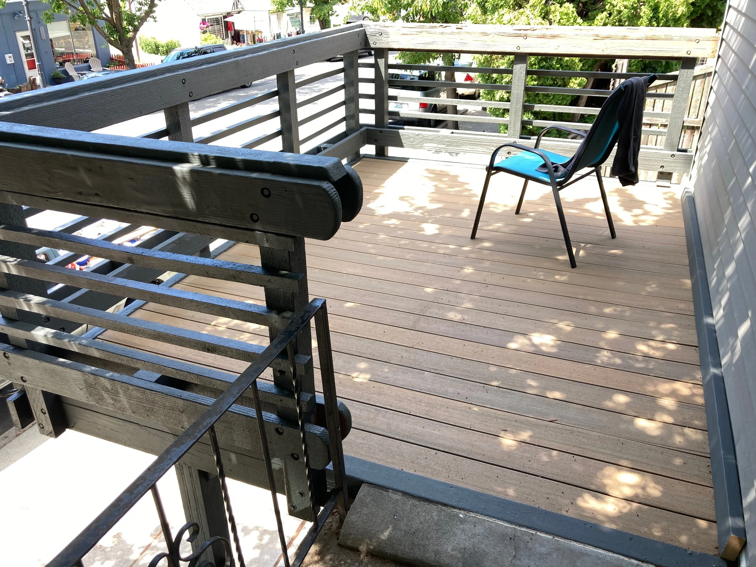 A wooden balcony with a black chair, a black jacket hanging over the chair's back, and shadows of leaves on the floor.