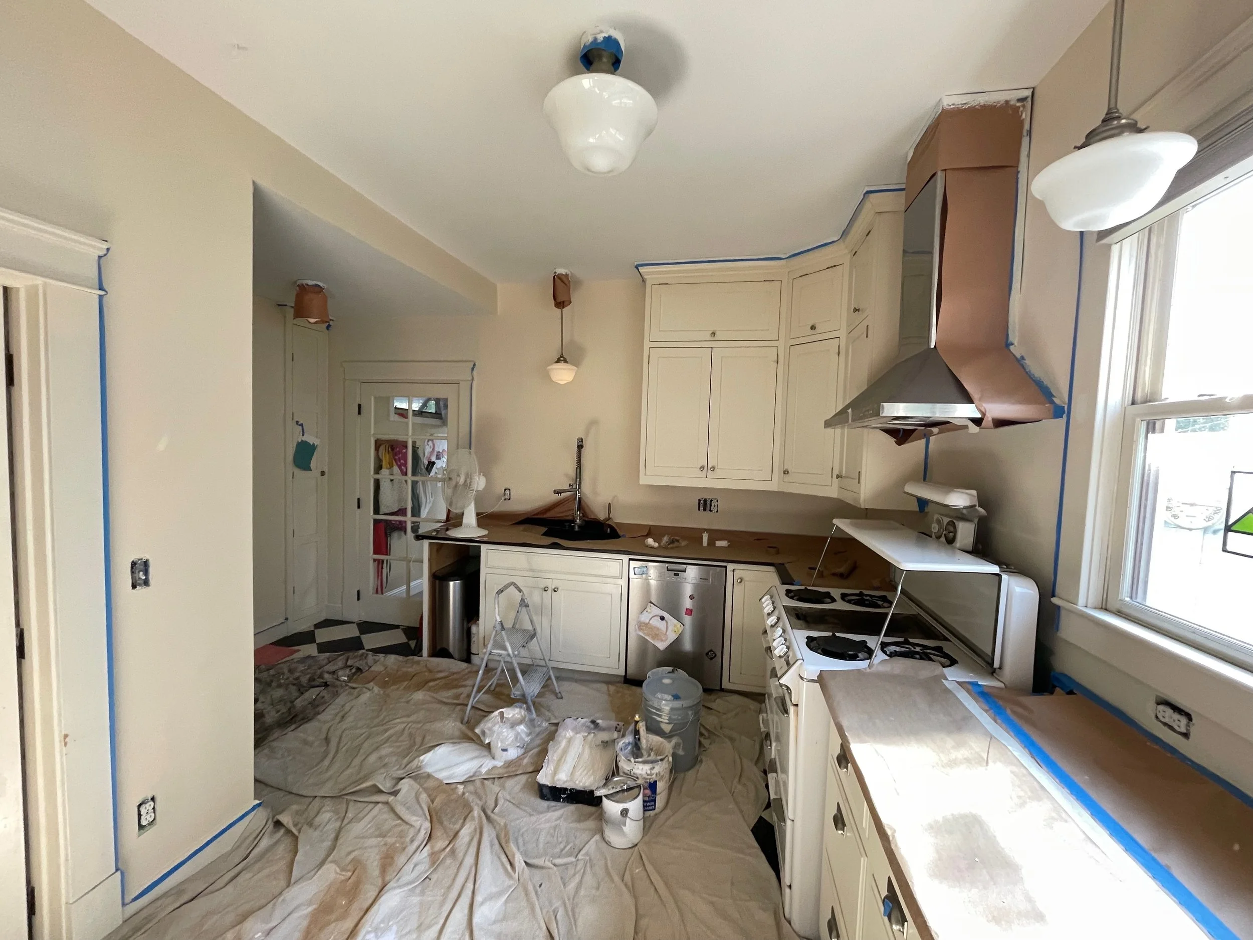 Kitchen undergoing renovation with painter's drop cloth on the floor, painting supplies, and partially painted areas on the walls and cabinets.