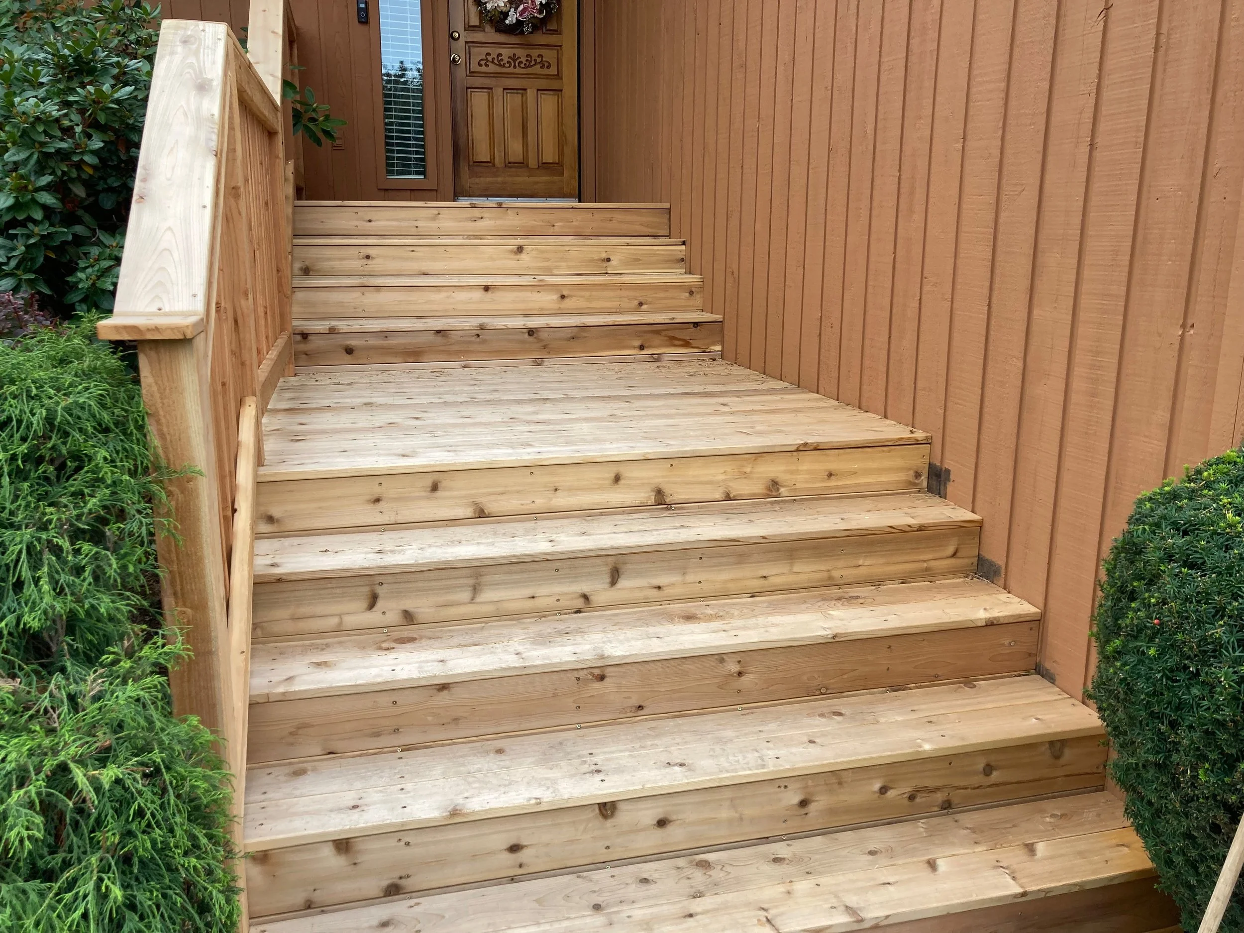 Newly built wooden front porch and steps leading up to a house with a brown door and window. Green bushes on either side of the stairs.