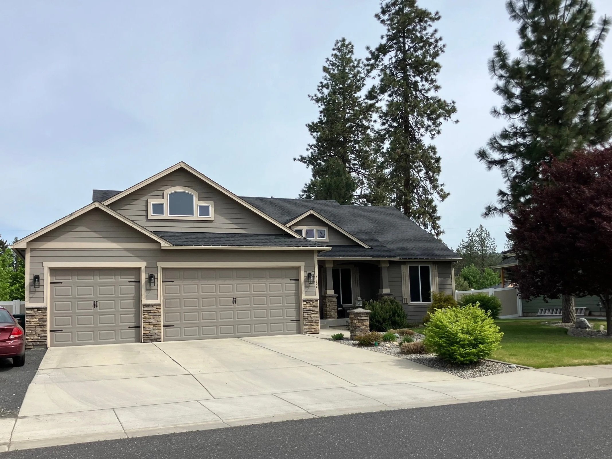 A modern, two-story house with beige siding, a dark shingle roof, and a three-car garage. The house has a small front porch, landscaped yard with shrubs and trees, and is situated on a paved driveway.