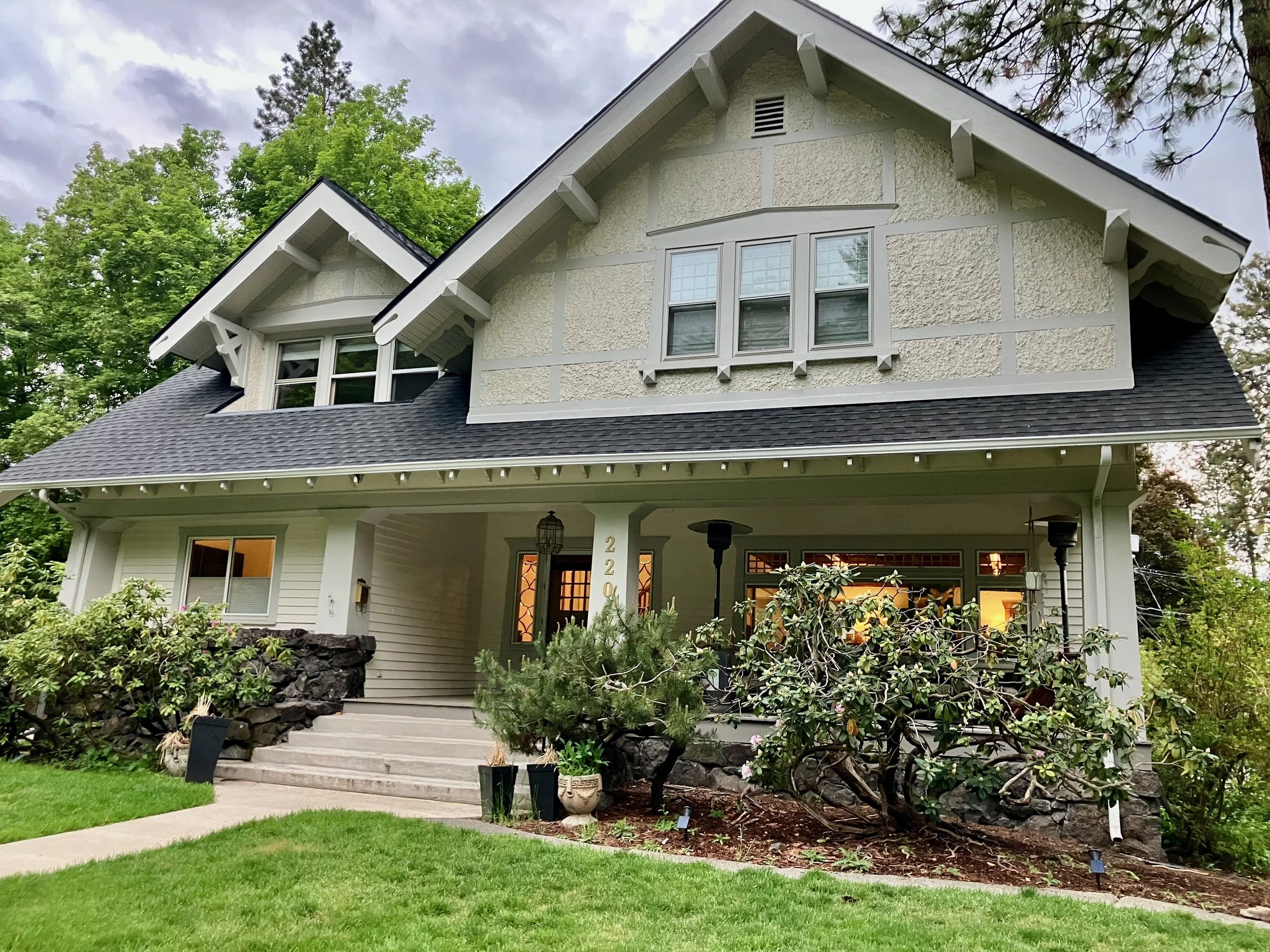 A two-story house with a gray roof, white siding, and beige stucco exterior, surrounded by greenery and trees.
