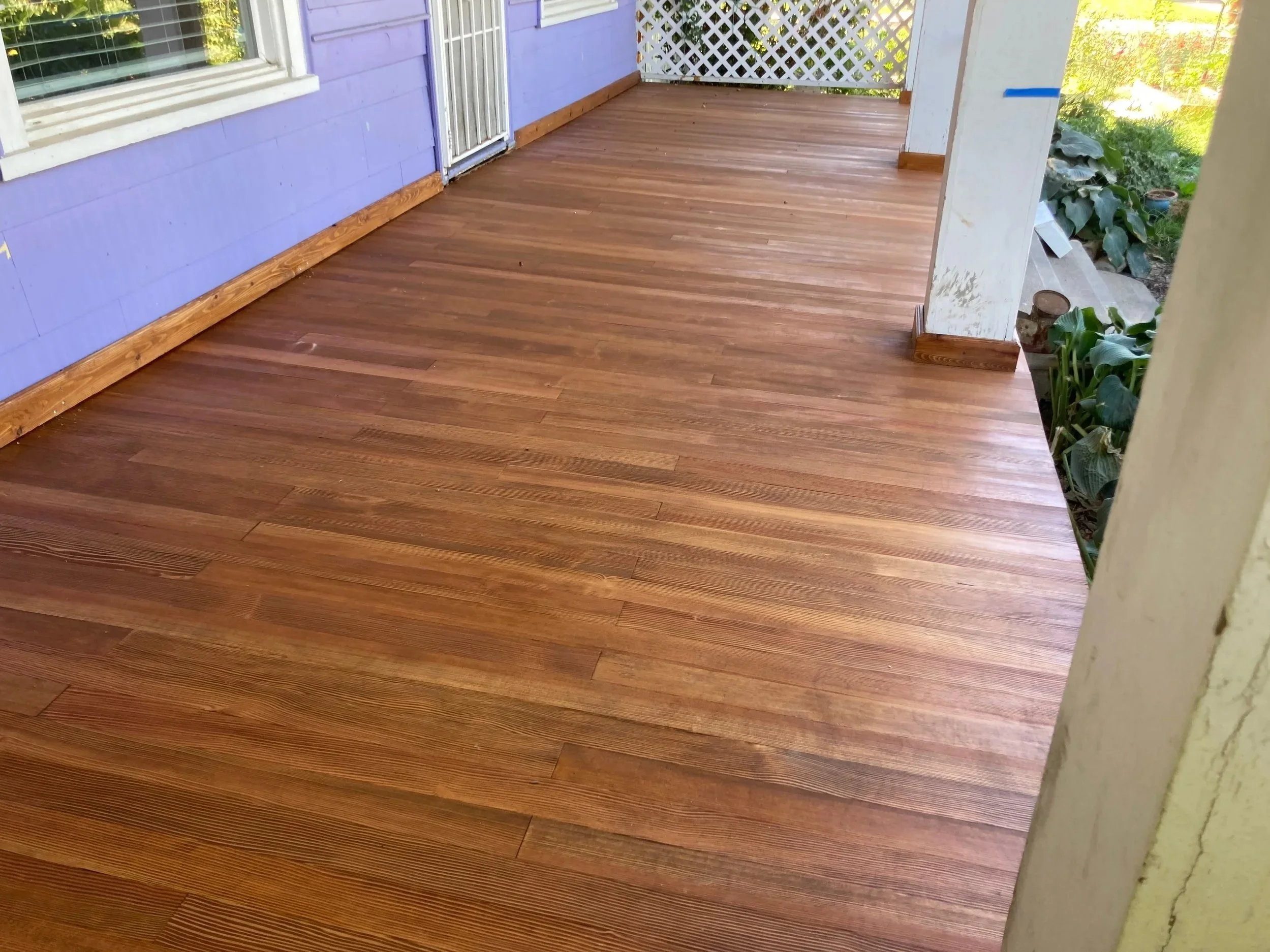 Freshly installed wooden porch flooring on a house with purple walls and a white lattice fence in the background.