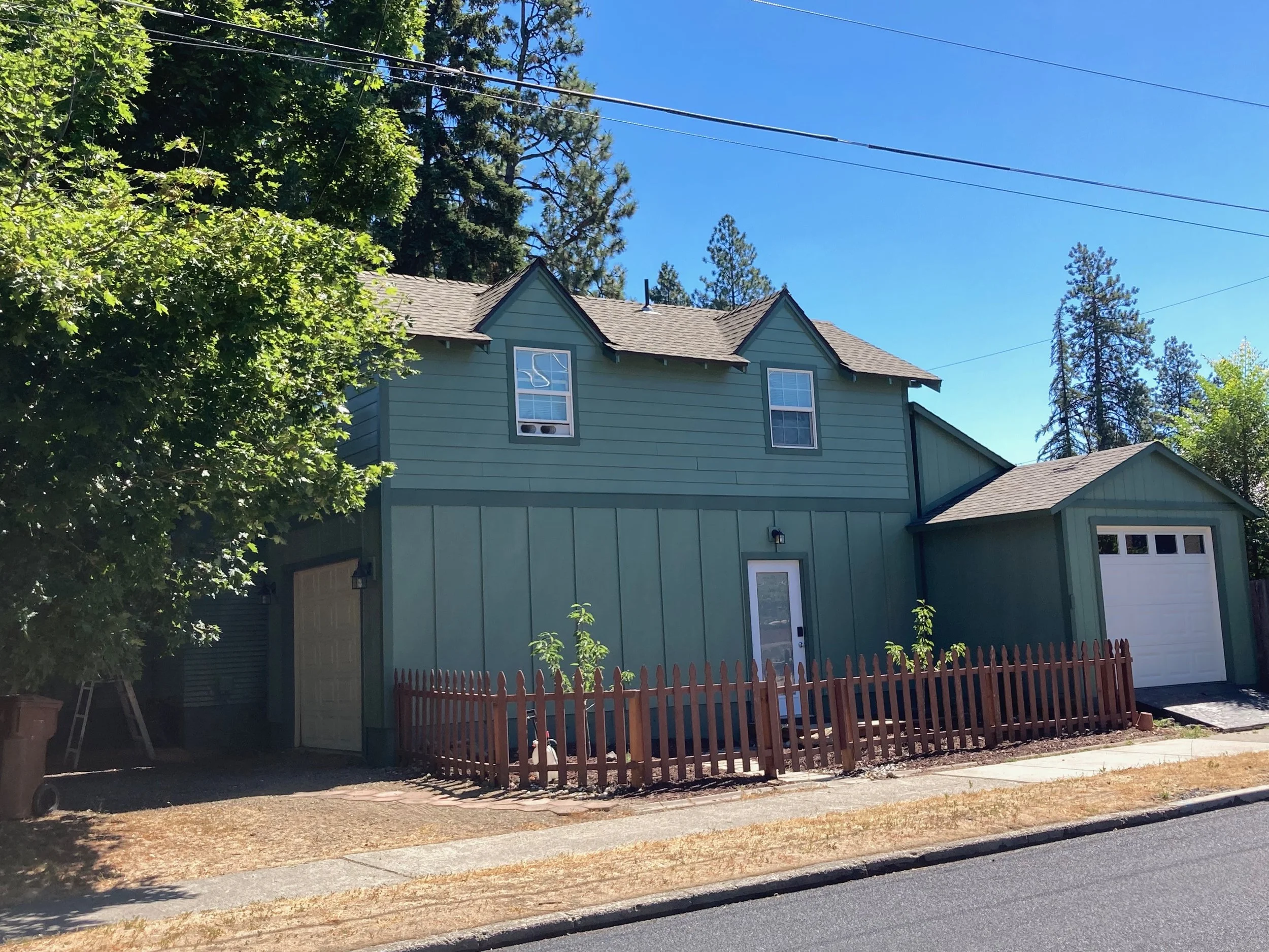 Green two-story house with white garage door, small fenced garden, and trees in front on a clear sunny day.
