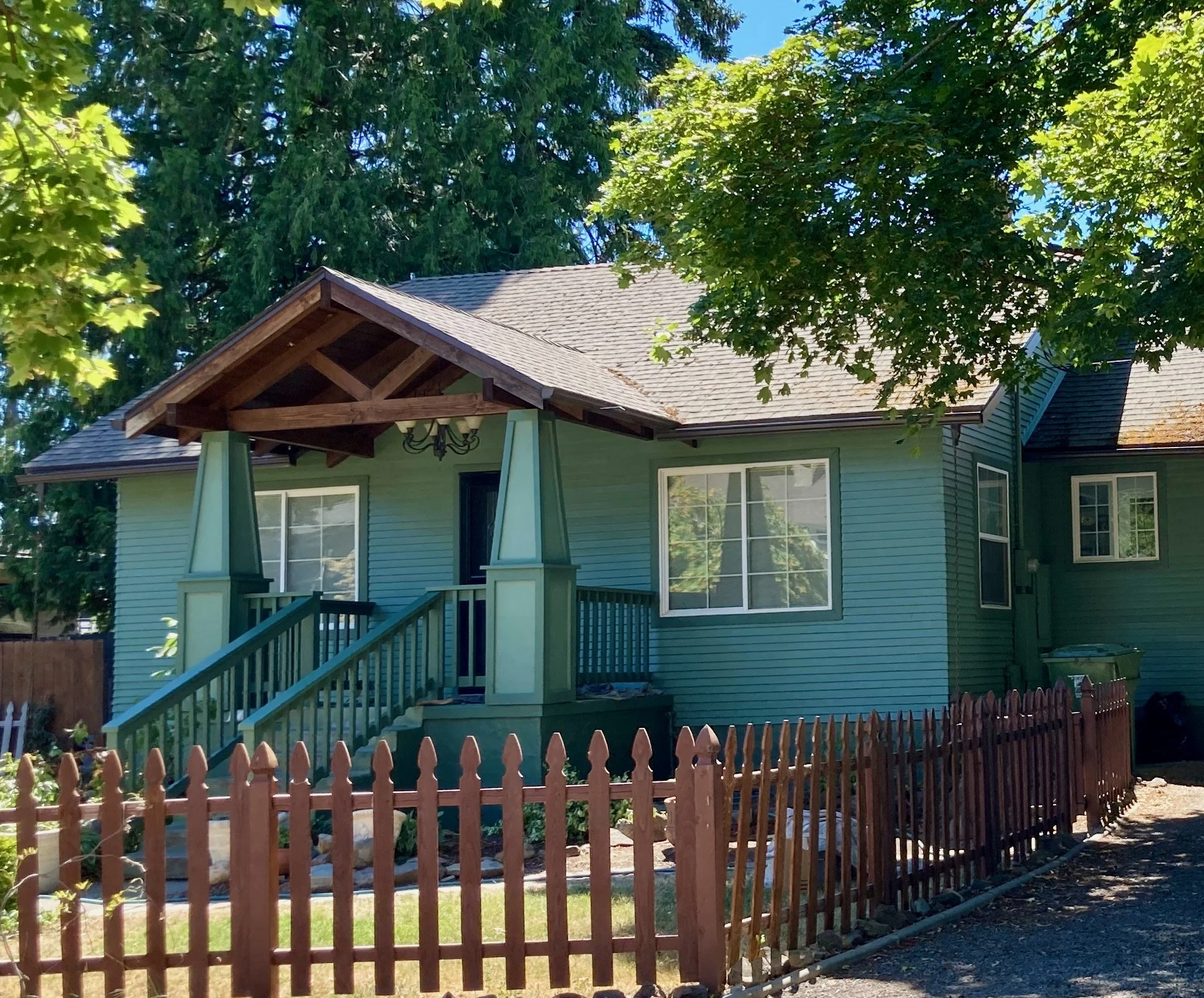 A blue house with a front porch, stairs, and a brown picket fence, surrounded by trees in a sunny outdoor setting.