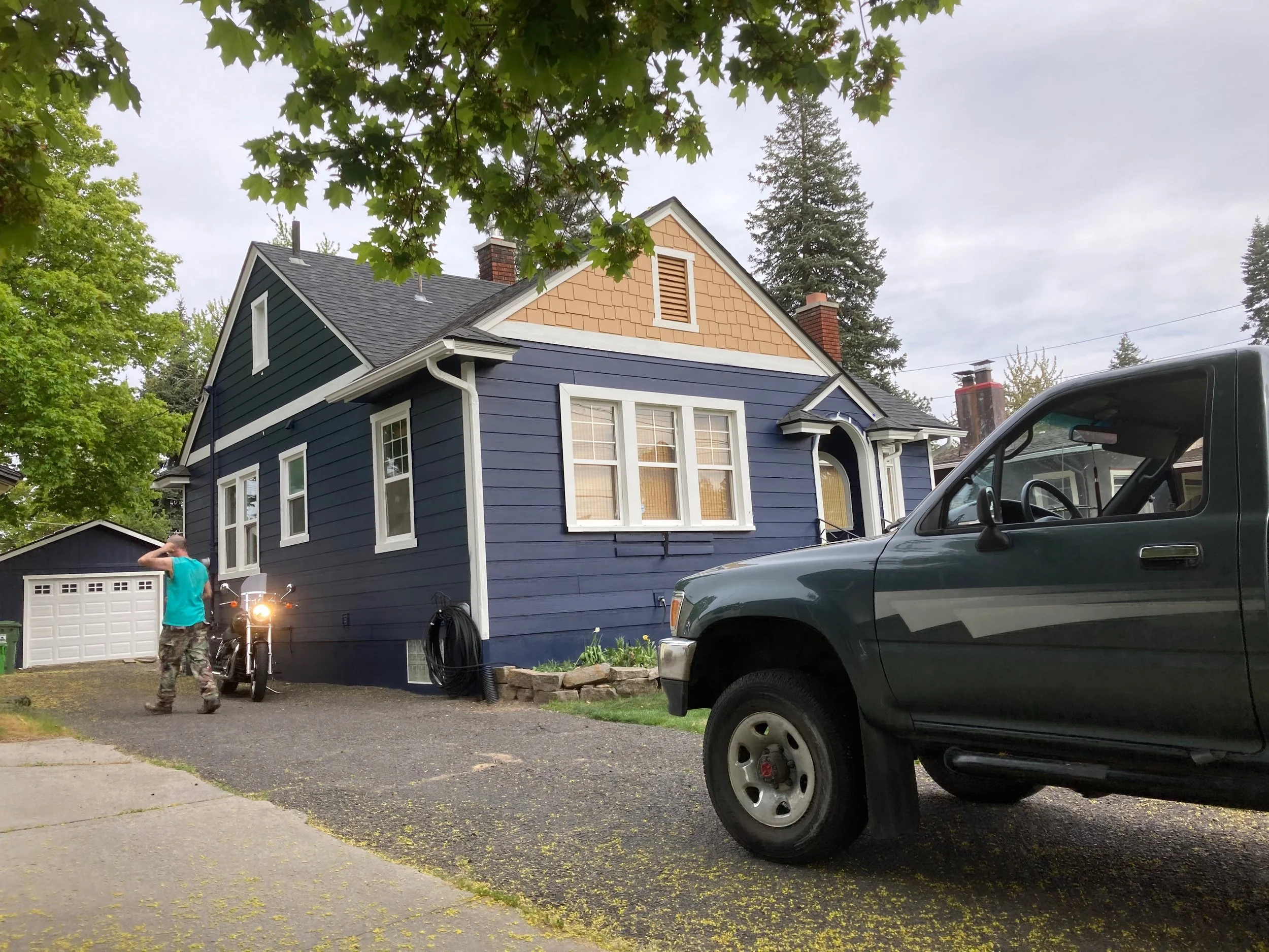 A blue house with a tan gable and multiple white-framed windows is shown. A man in a teal shirt and camo pants is standing near a motorcycle, talking on the phone. A green pickup truck is parked in the foreground on the right side, and there is a garage in the background. The scene is outdoors with trees and a cloudy sky.
