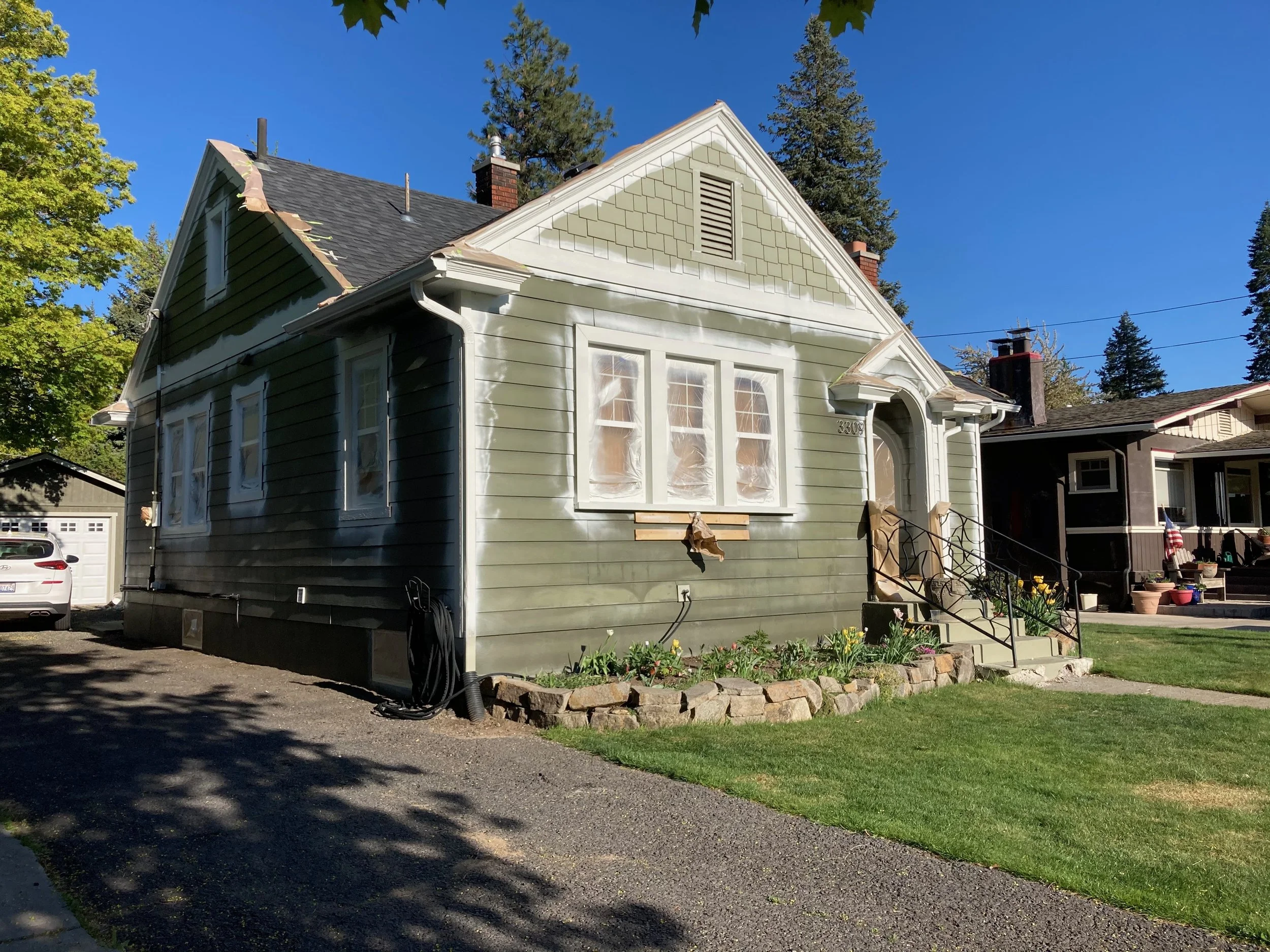 A house undergoing exterior renovation, with windows covered in plastic, green siding, and a small front yard with flowers surrounded by stones.