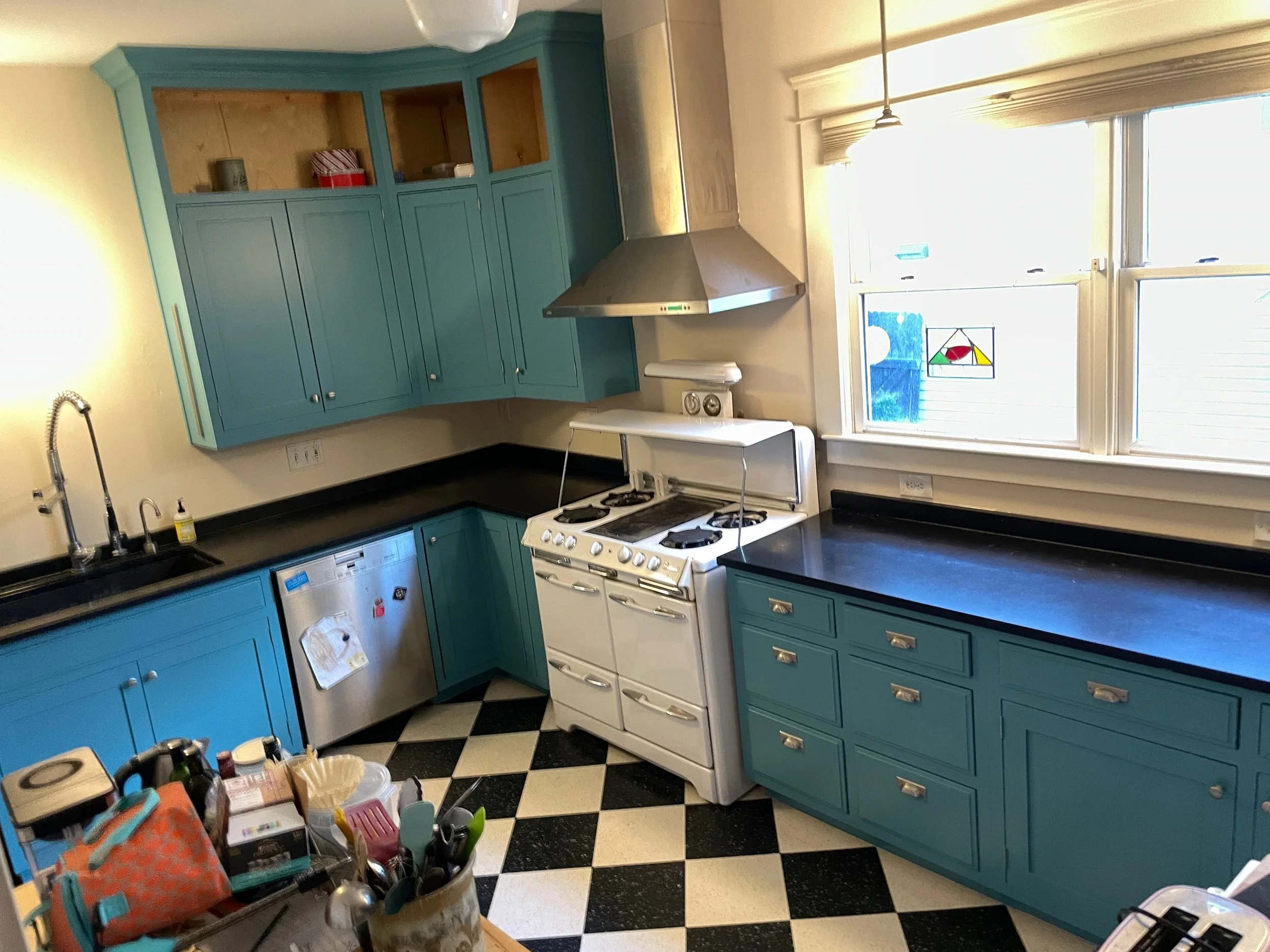 A kitchen with teal cabinets, a black countertop, a white stove, and black and white checkered flooring. There is a window with stained glass, and clutter on the counter and a table.