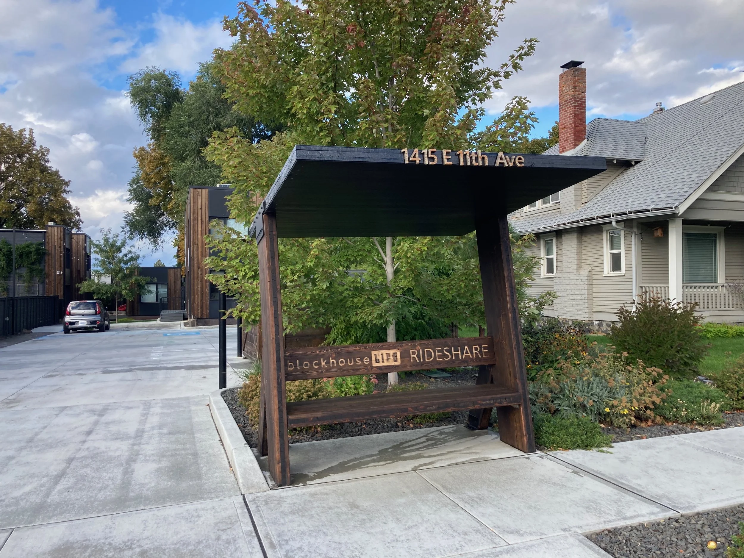 A wooden bus stop shelter with a sign displaying the address 1415 E 11th Ave. The shelter has a sign underneath that reads 'blockhouse rideshare.' There's a green tree behind the shelter, with a house, parked cars, and a modern building in the background during a cloudy day.