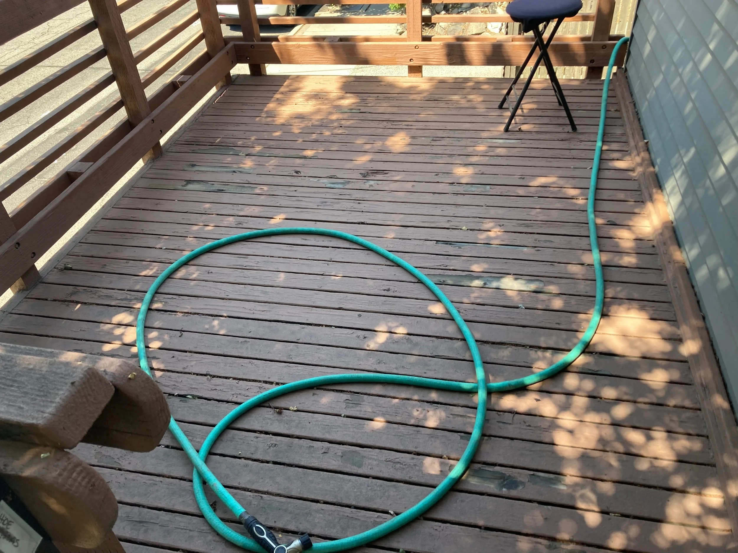 A wooden deck with a garden hose coiled on the floor and a black folding chair against the wall, with shadows of leaves on the deck