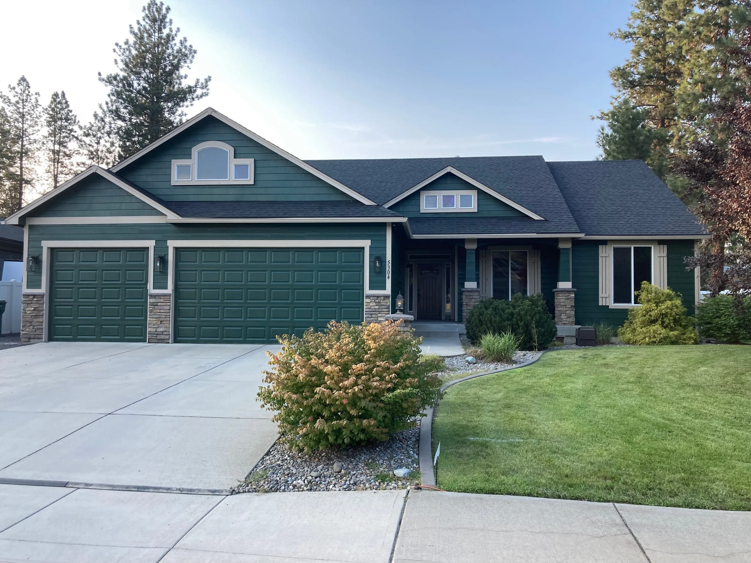 Front yard of a large house with green siding, stone accents, three-car garage, and a well-maintained lawn with bushes and trees.
