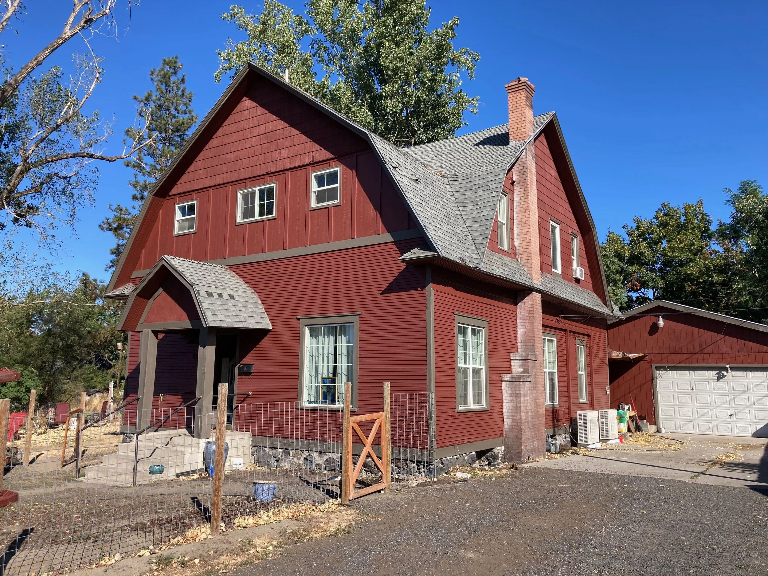 A red, two-story house with a steep gabled roof and multiple windows. The house has a small covered entrance with steps, a brick chimney, and a fenced yard. There are trees and a blue sky in the background.