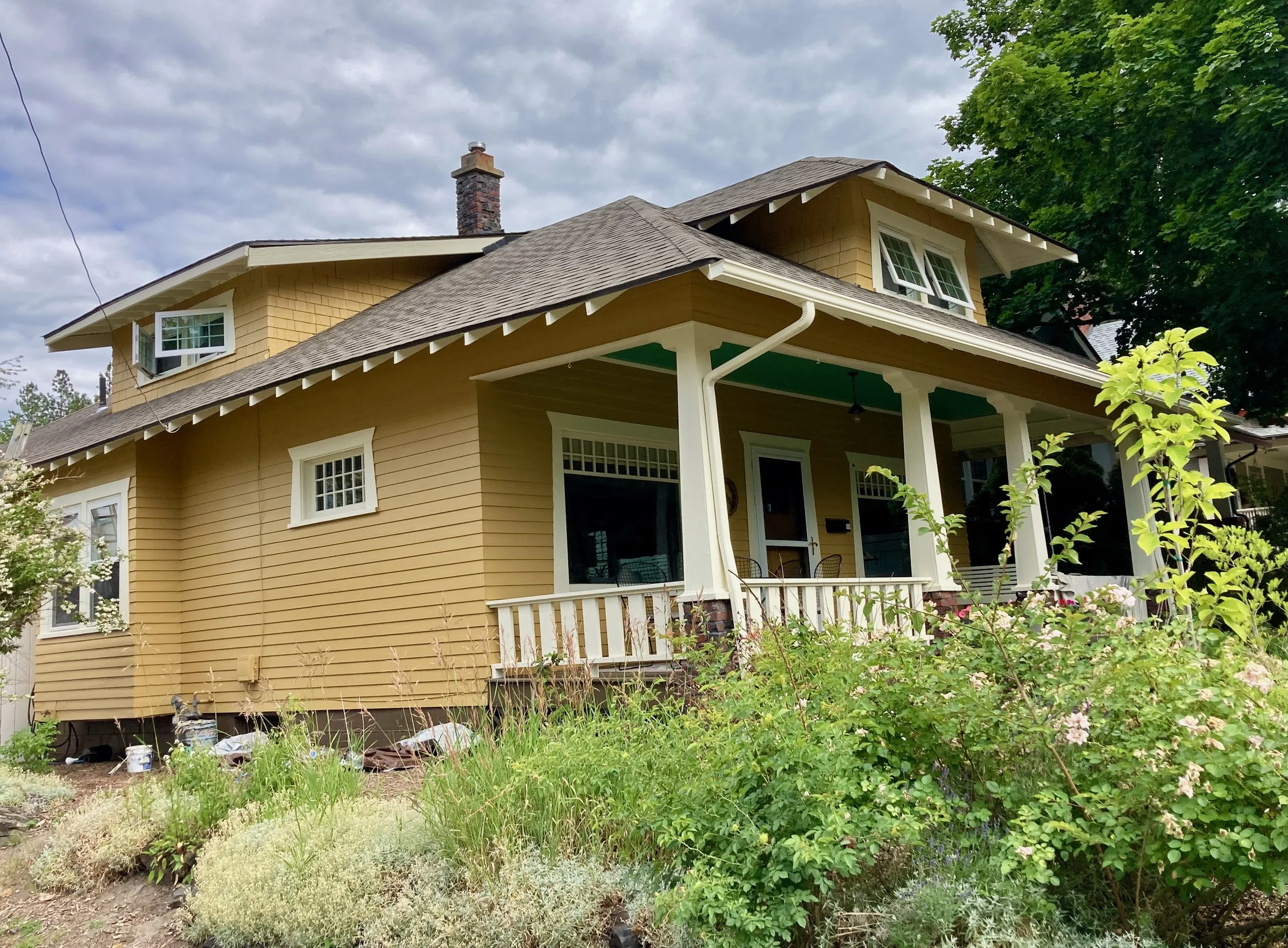 Yellow two-story house with a porch, multiple windows, and a chimney, surrounded by green shrubs and trees on a cloudy day.