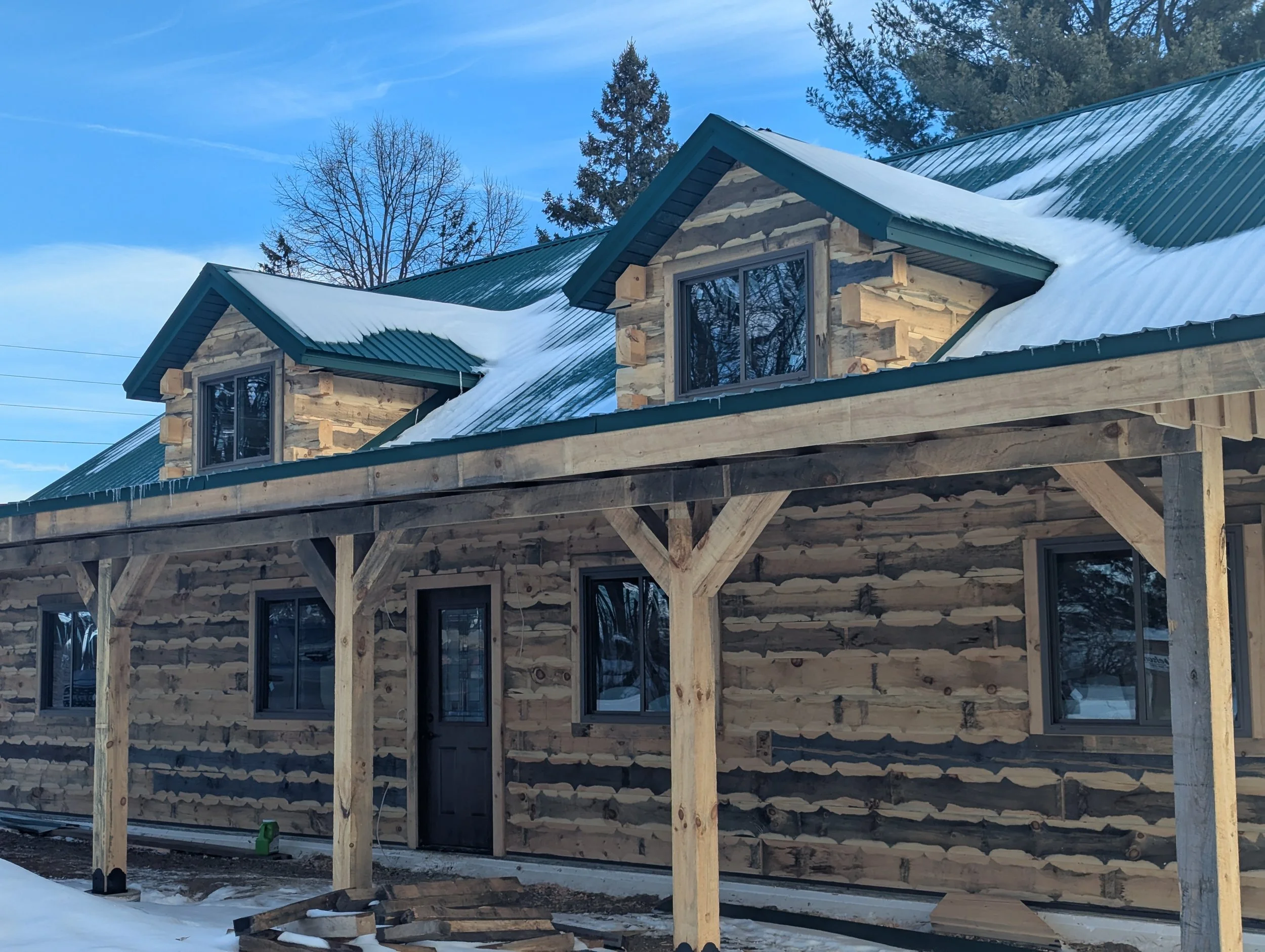 A partly constructed log cabin with a green metal roof and snow on top, surrounded by trees and blue sky.