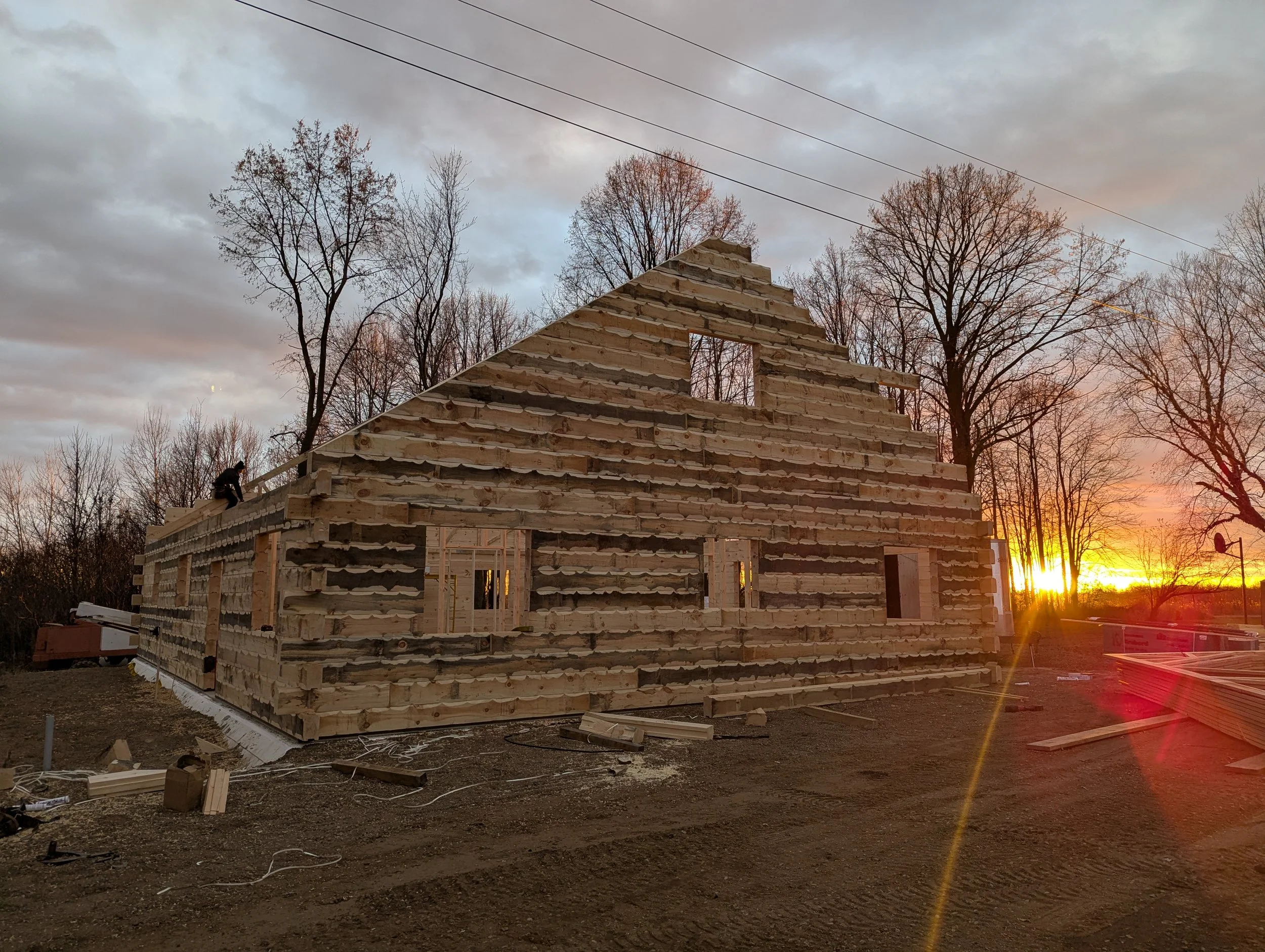 A house under construction made of wooden logs, with front and side openings for windows and doors, at sunset with a partly cloudy sky and leafless trees in the background.