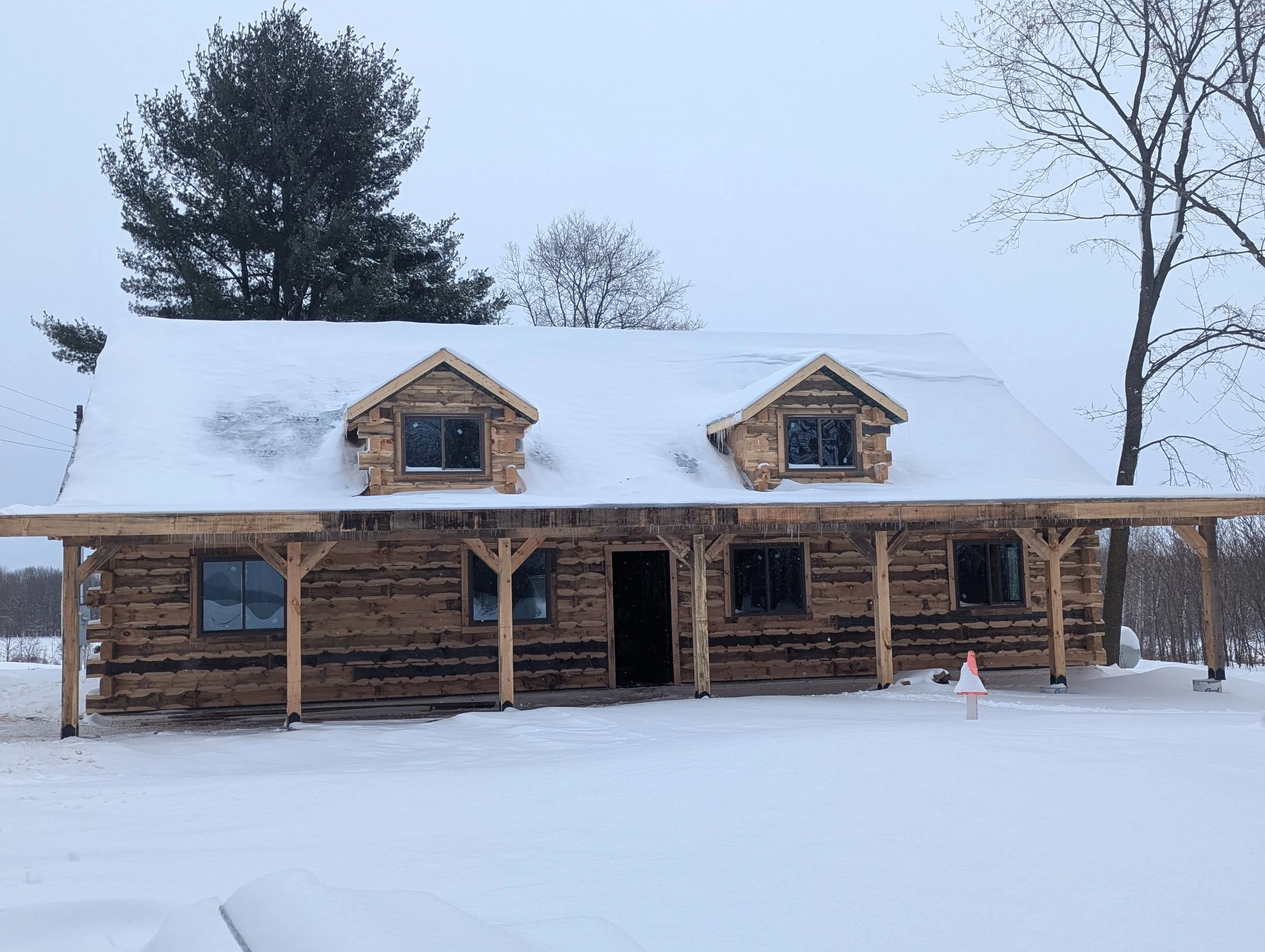 A two-story log cabin under construction on a snowy landscape, with a large snow-covered roof and a porch supported by wooden posts, surrounded by leafless trees and a cloudy sky.