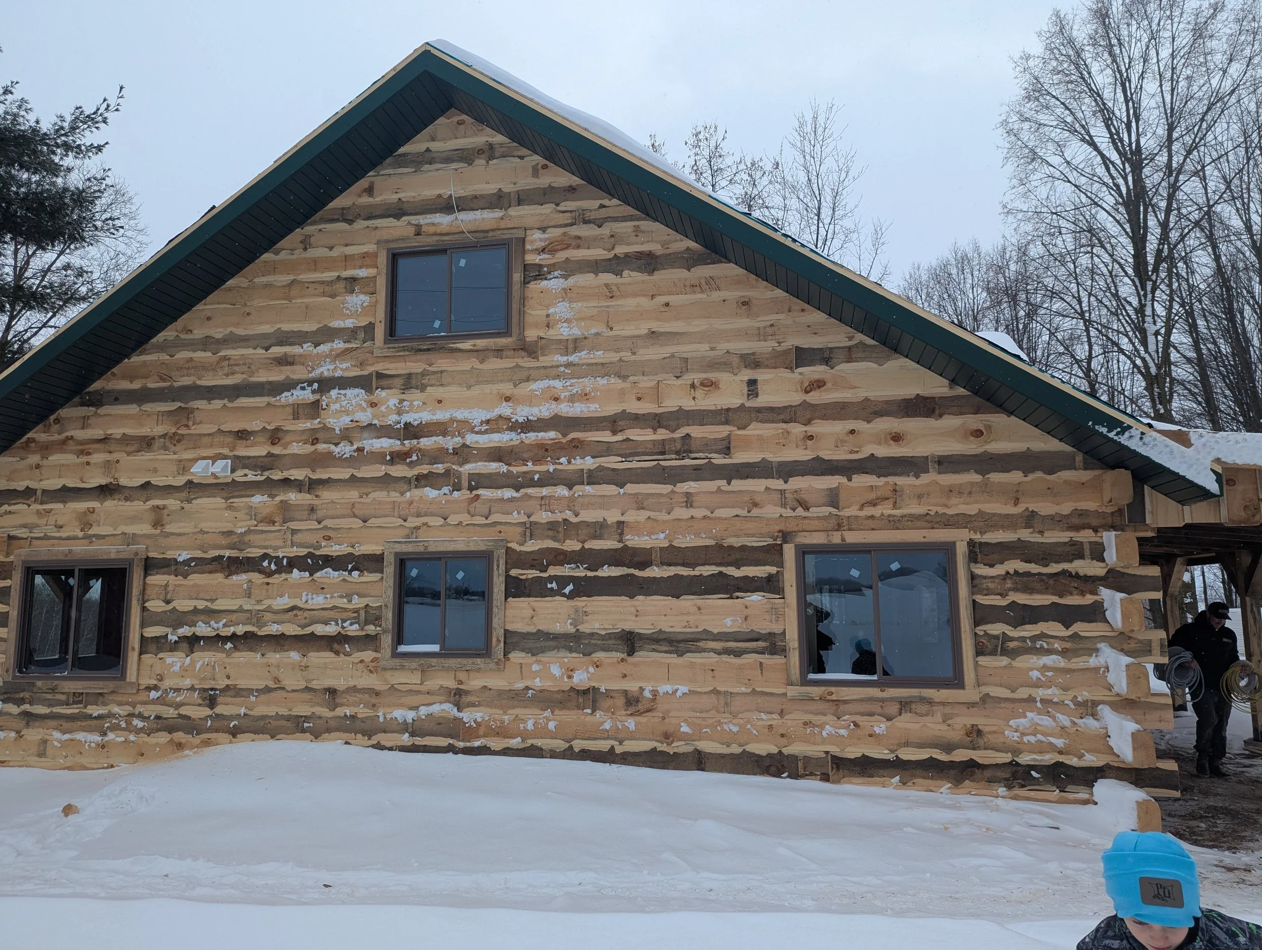 A partially constructed wooden house in a snowy landscape, with some construction workers visible on the right side.
