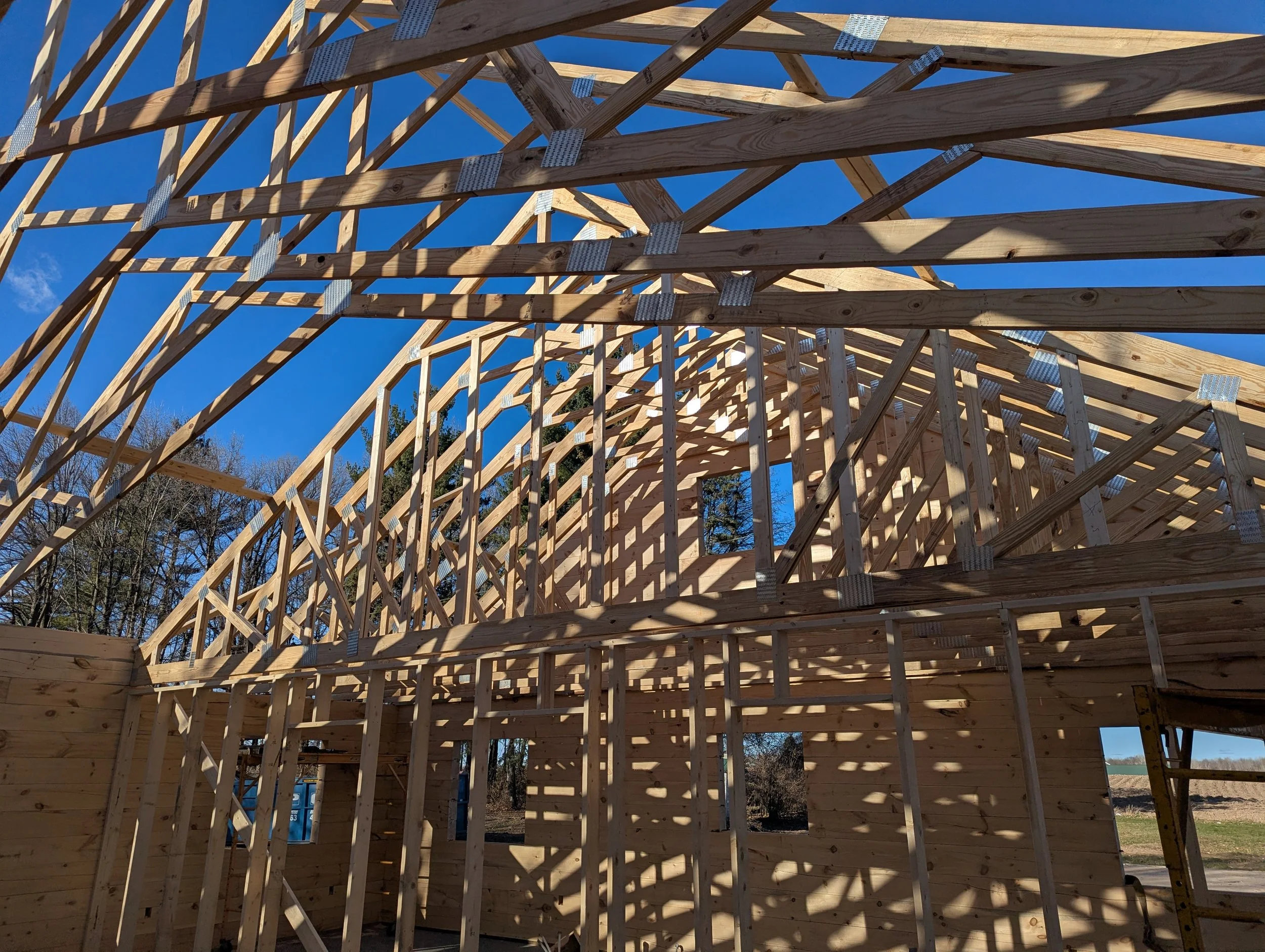 Wooden framing of a building under construction with shadows cast by the framework on the walls and roof, and trees visible in the background under a blue sky.