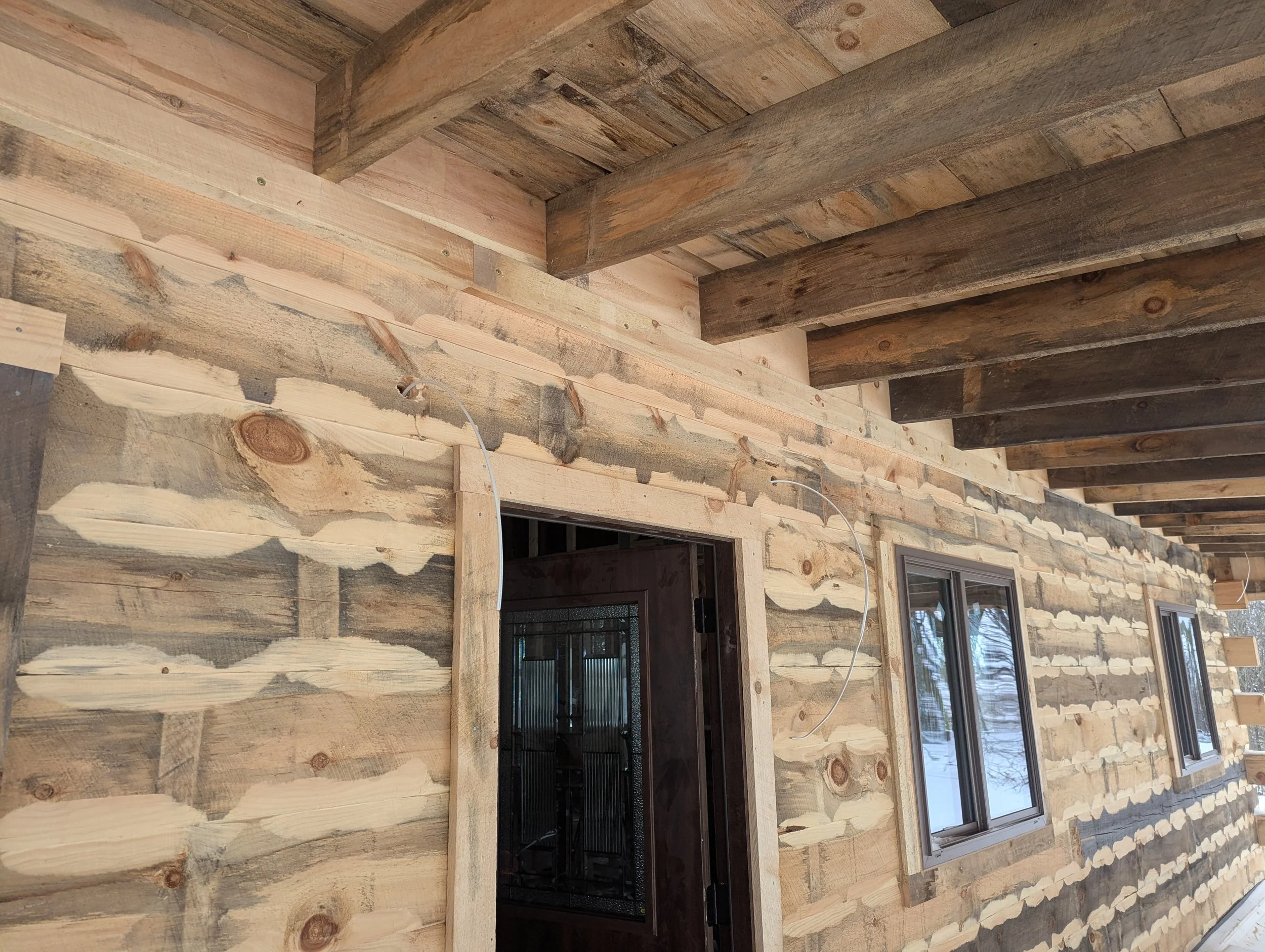 Wooden house exterior under construction with stained and unstained wood siding, window frames, and ceiling beams.