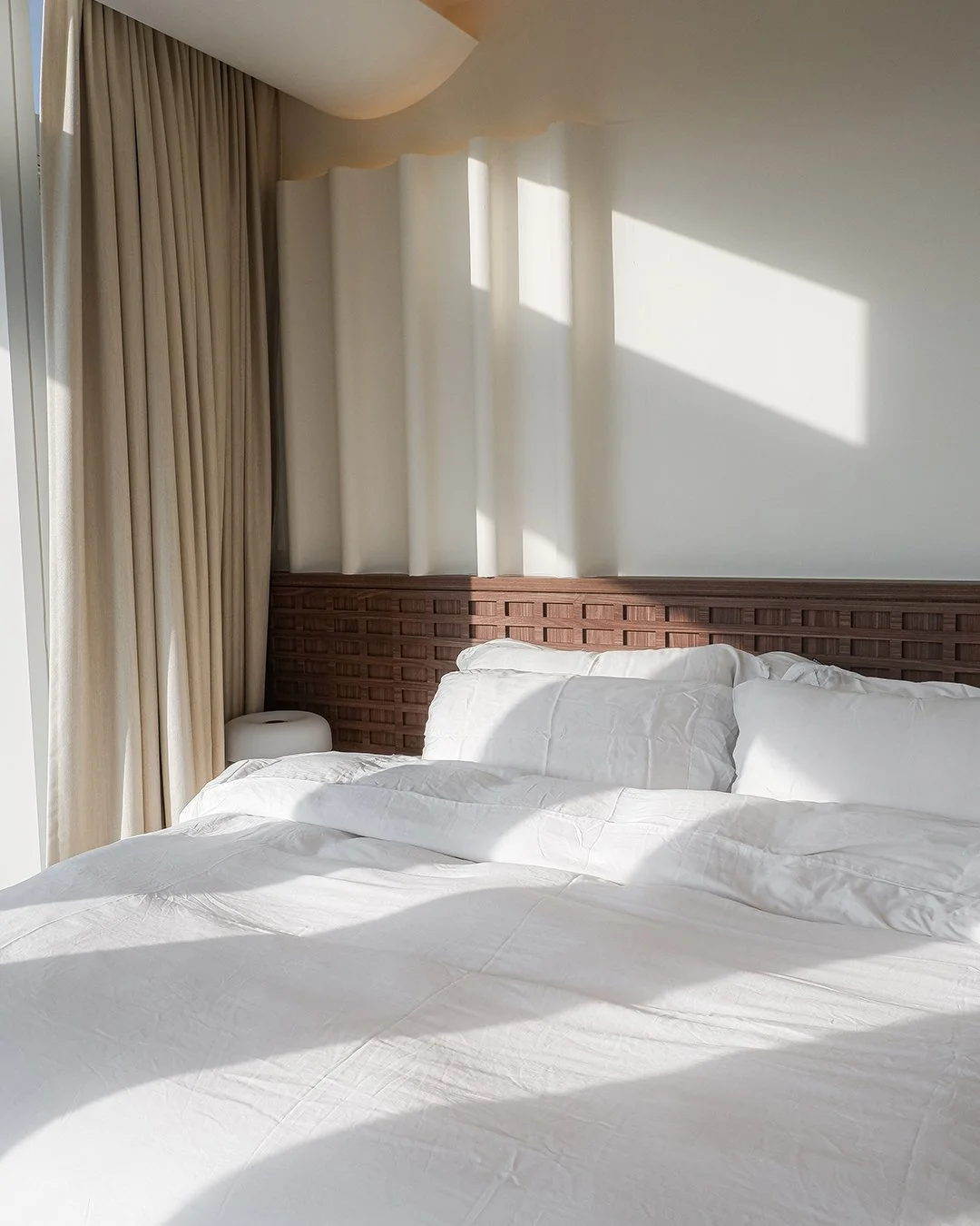Sunlit bedroom with a white bed, pillows, beige curtains, and a wooden headboard, casting shadows on the wall.