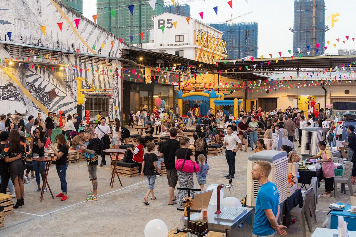 People gathered at an outdoor event with colorful decorations, balloons, and a bounce house, surrounded by buildings.