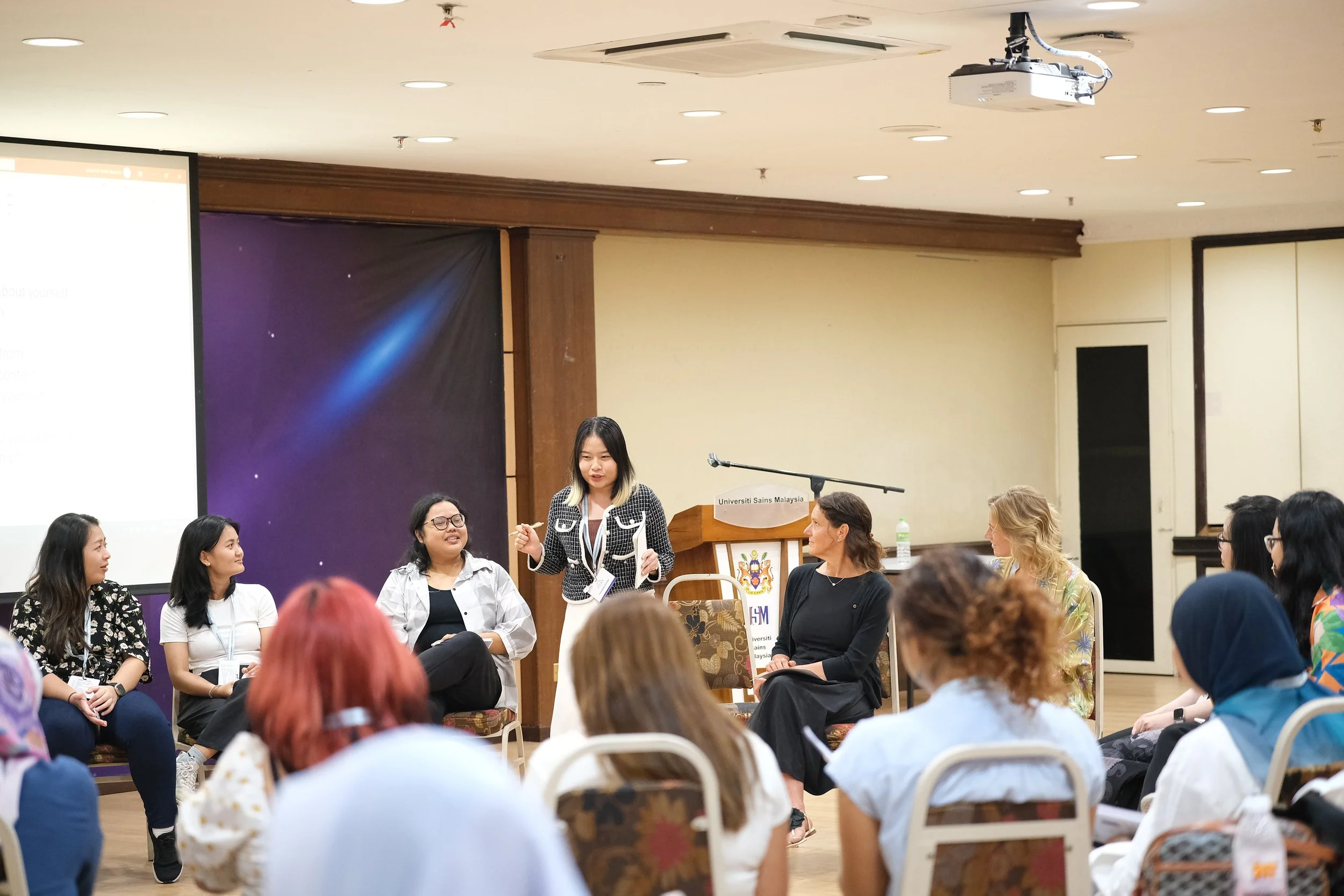 A woman standing and speaking in front of an audience during a panel or workshop in a conference room at Universiti Sains Malaysia. Several women are seated and listening to her.