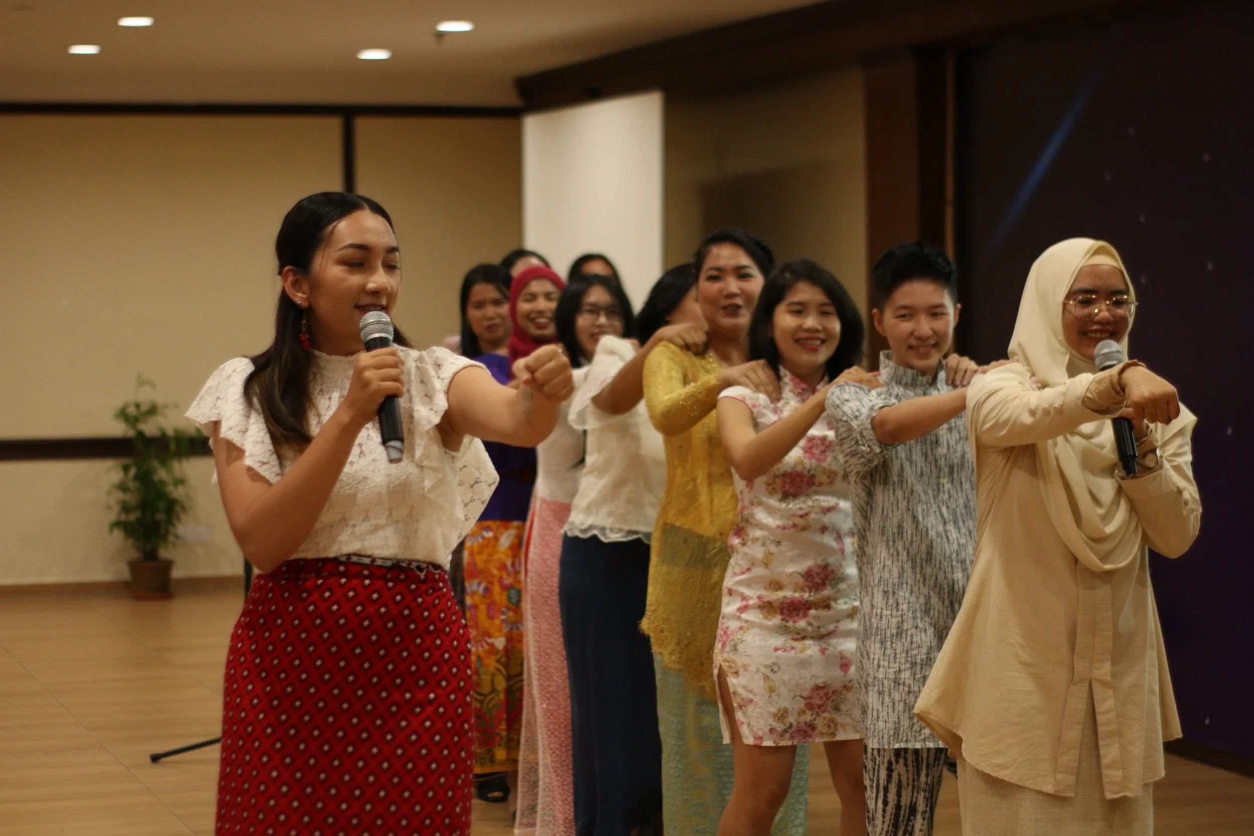 Group of women standing in a line, holding microphones, with their arms on each other's shoulders, engaging in a team-building activity or performance in an indoor setting.