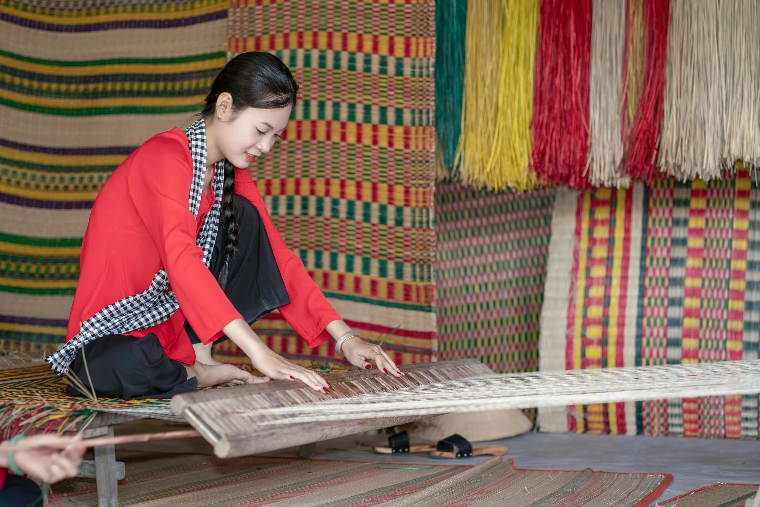 A woman in a red jacket and black pants scrapes a traditional woven textile on a wooden loom, with colorful woven wall hangings behind her.
