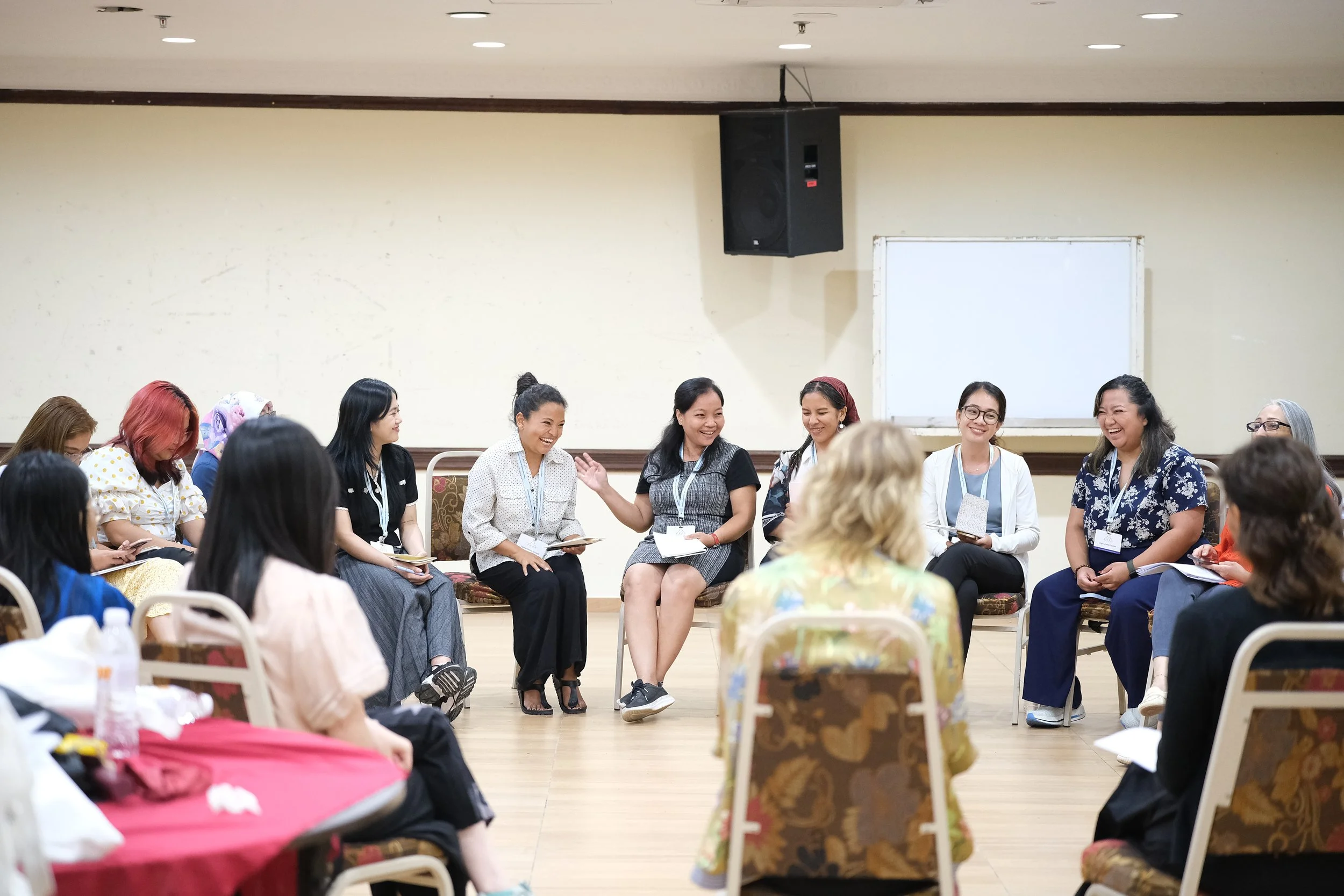 Group of women seated in a circle participating in a discussion or workshop, with some women smiling and engaging with each other in a room with a blank whiteboard in the background.