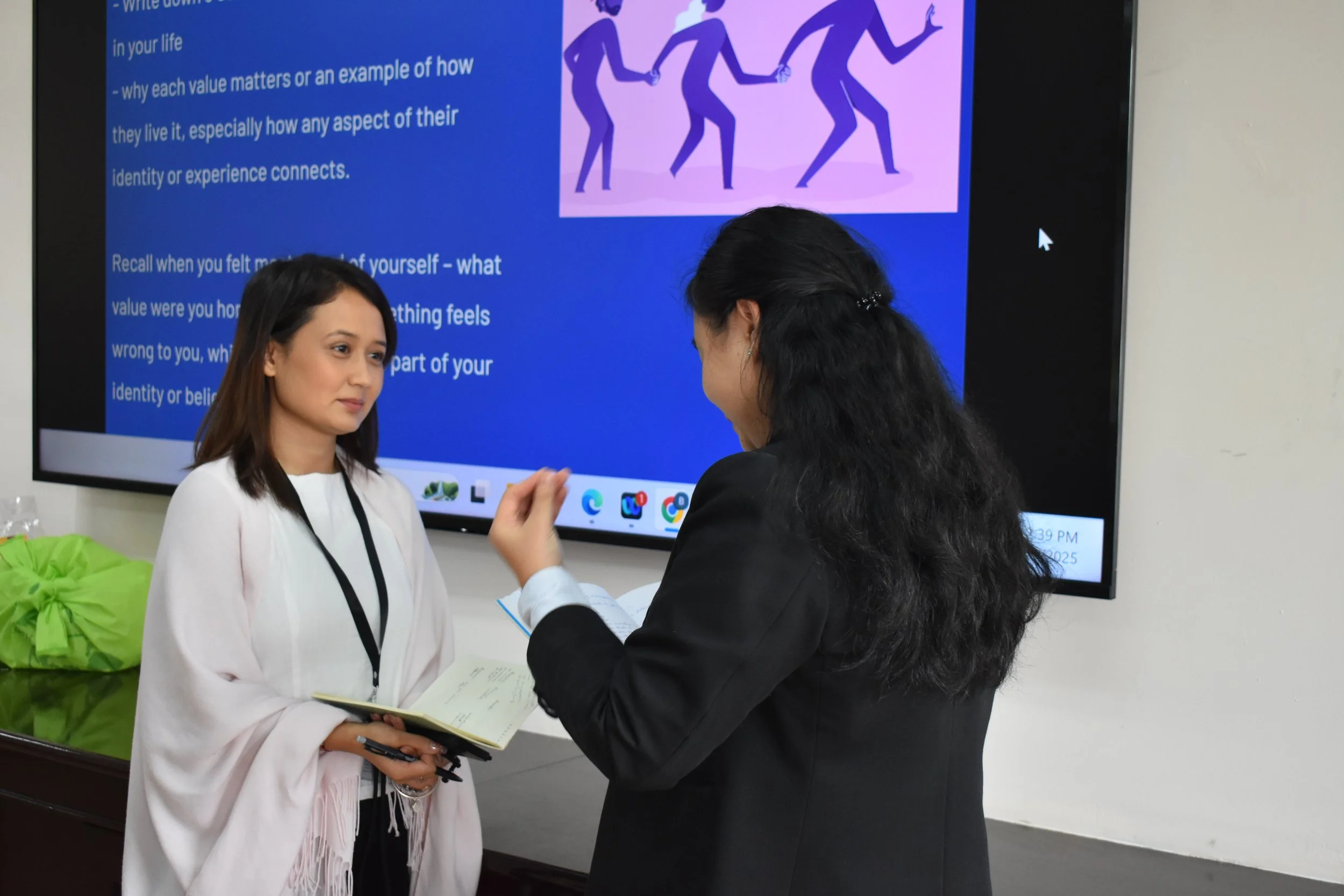 Two women having a discussion in front of a presentation screen. One woman is holding a notebook and pen, the other is speaking and gesturing with her hand. The presentation screen displays text and an illustration of people dancing or exercising.