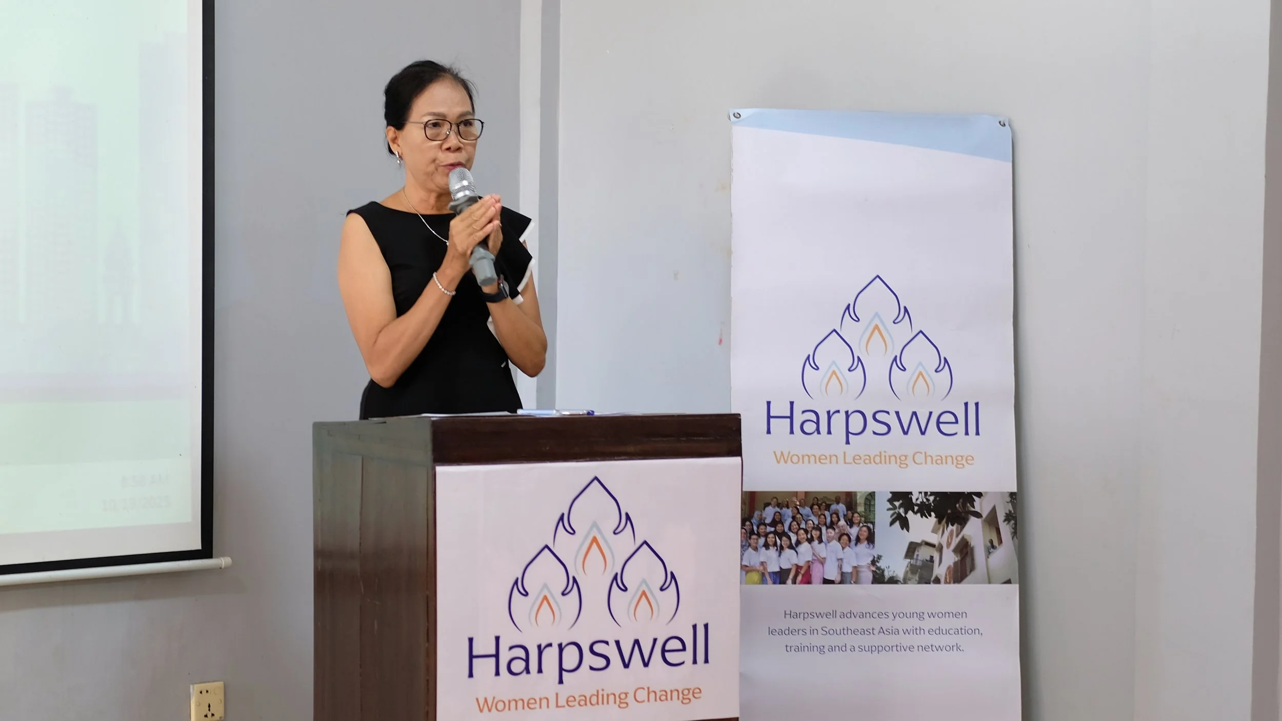 A woman in black dress with glasses speaking into a microphone behind a podium with a Harpswell banner, which features a logo of leaves and the tagline "Women Leading Change," at a conference or event.