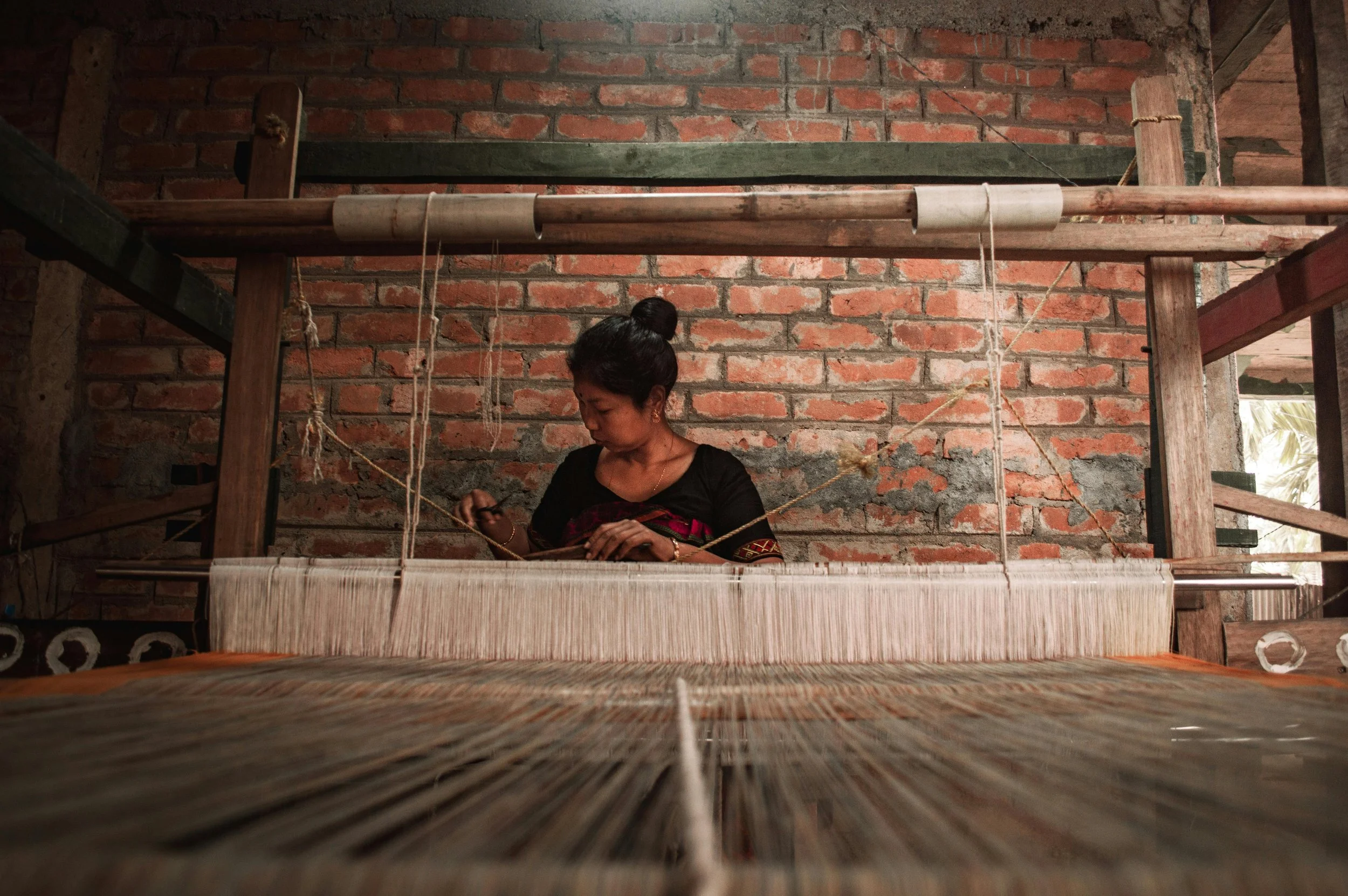 A woman weaving fabric on a traditional loom against a brick wall.