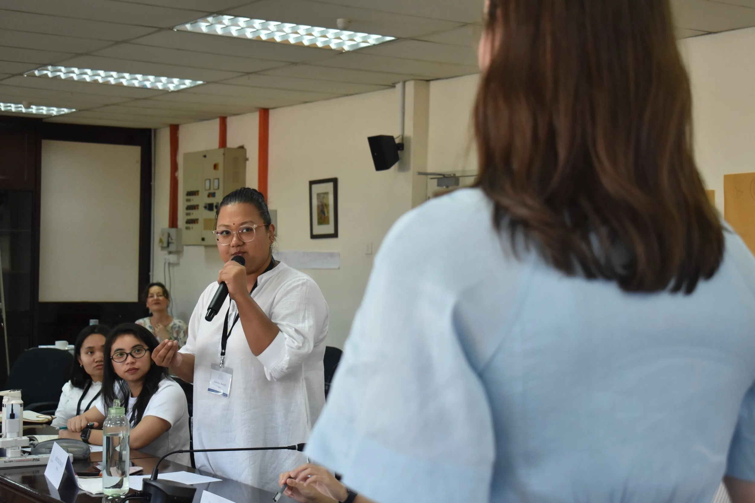 A woman wearing glasses and a white shirt is speaking into a microphone at a meeting or conference, with several other women sitting at a table in front of her, some looking at her attentively.