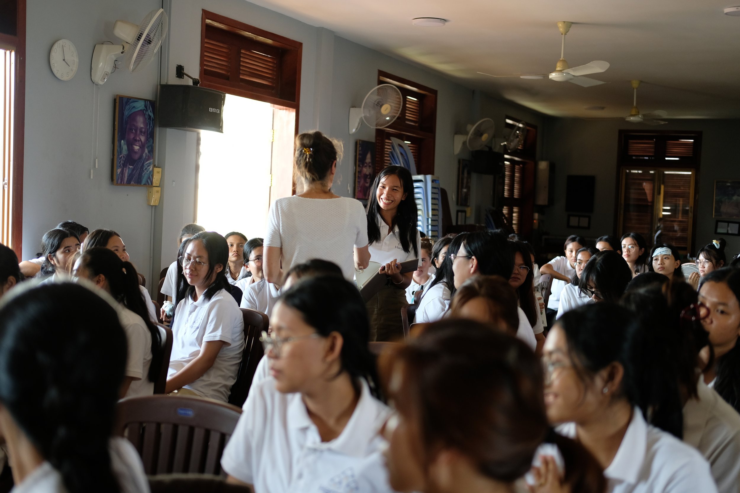 A large group of women, mostly in white shirts, seated in a room with wooden window shutters, smiling and engaging with each other. Two women are standing and talking, one holding a tablet, in a well-lit room with ceiling fans and portraits on the wall.