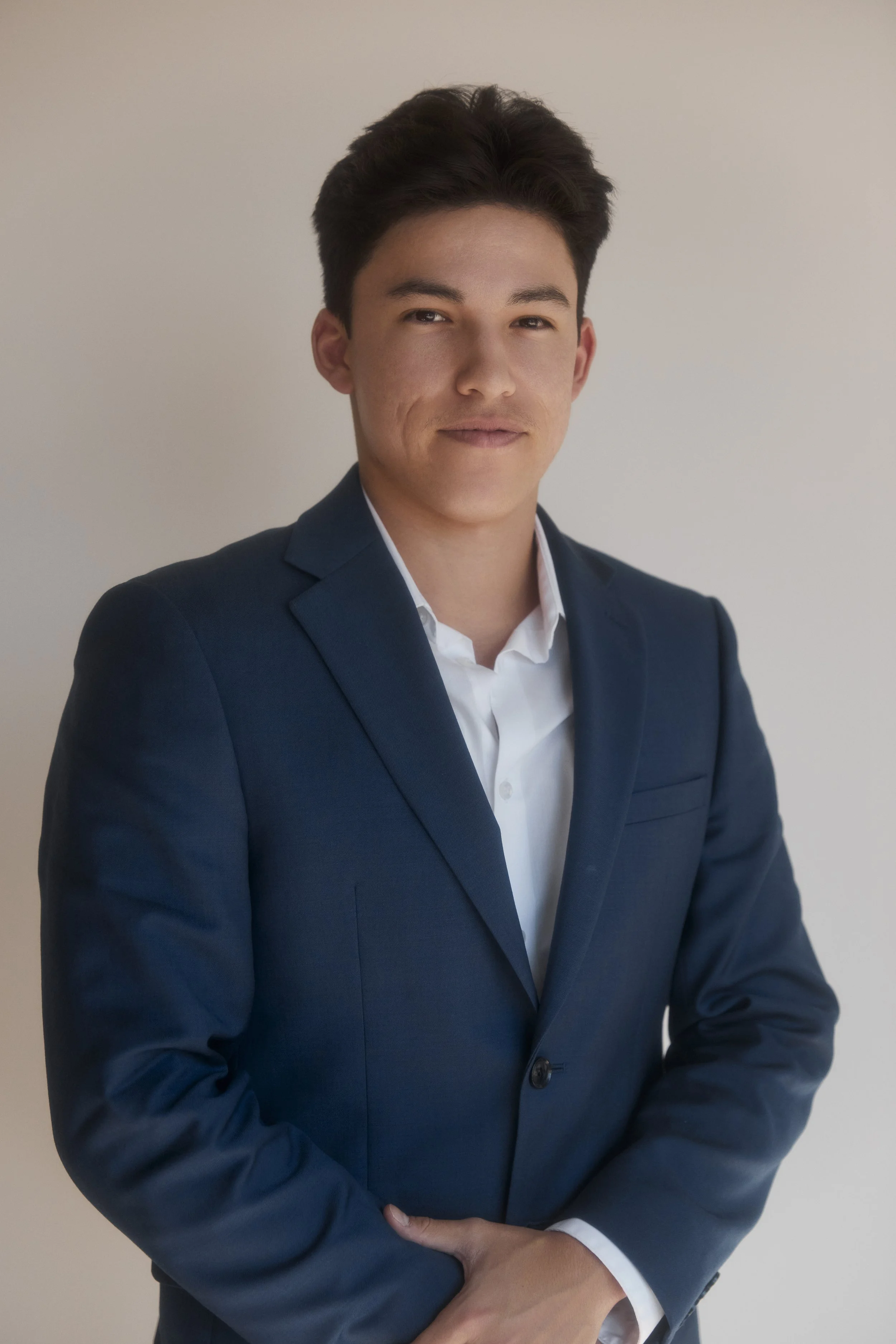 Young man wearing a dark blue suit and white dress shirt, standing against a plain light-colored background, looking at the camera with a slight smile.