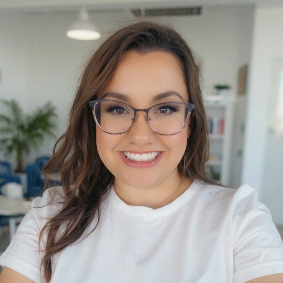 A young woman with long brown hair, glasses, and a white shirt smiling at the camera in an indoor setting with shelves and plants in the background.